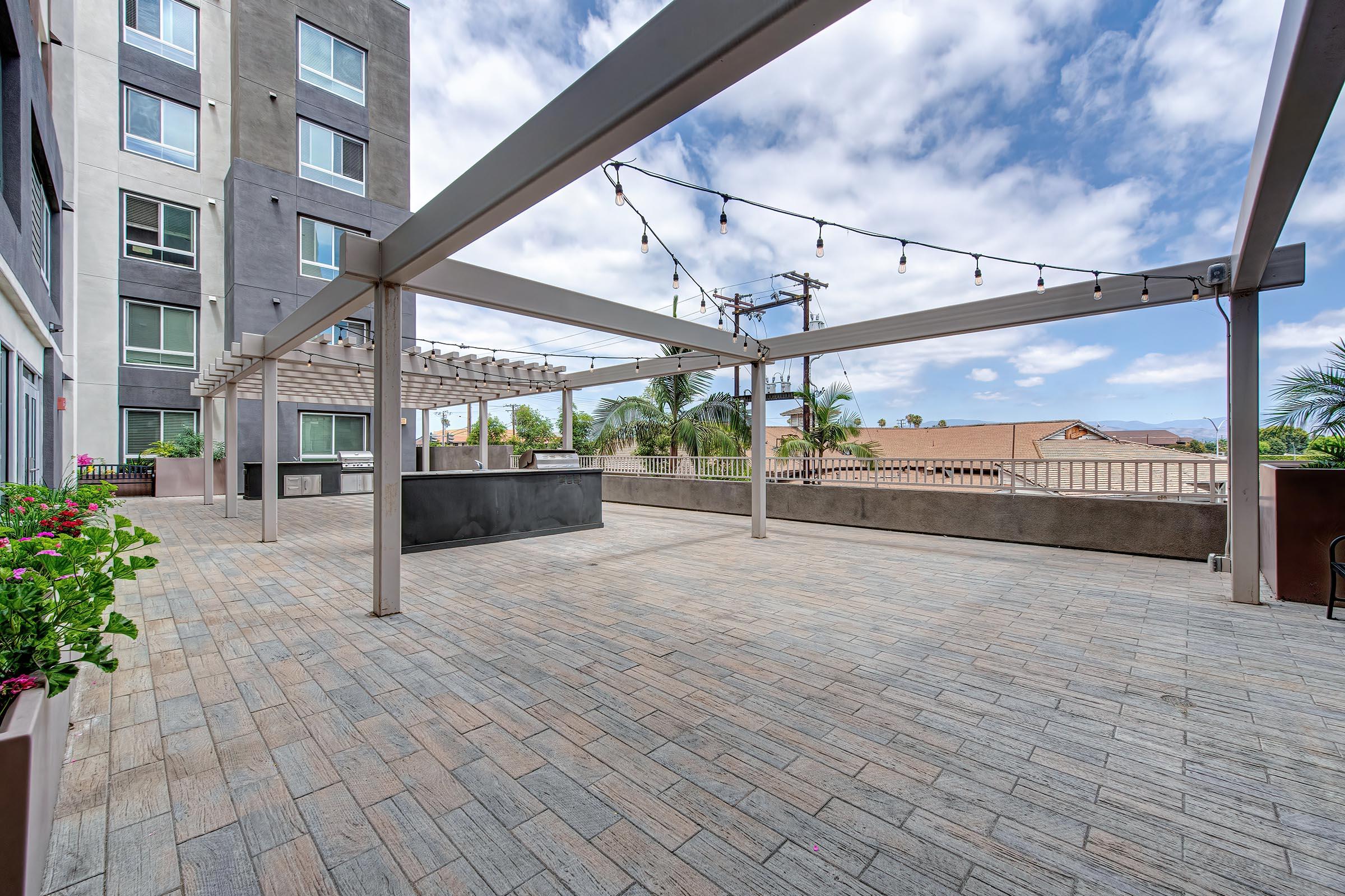 A spacious outdoor patio area featuring a pergola with string lights, surrounded by potted plants. The flooring is patterned stone, and the background includes a view of buildings and palm trees under a partly cloudy sky.