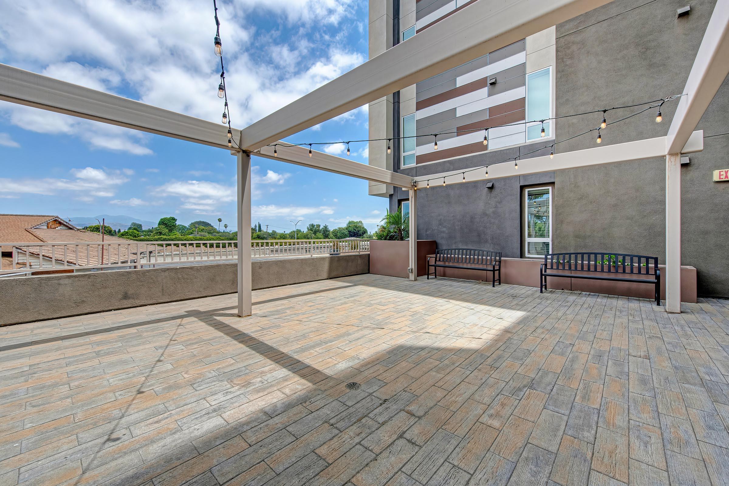 A spacious outdoor patio area featuring a paved floor, overhead string lights, and two benches against a wall. The sky is partially cloudy, and surrounding greenery is visible in the background, with a modern building adjacent to the terrace.