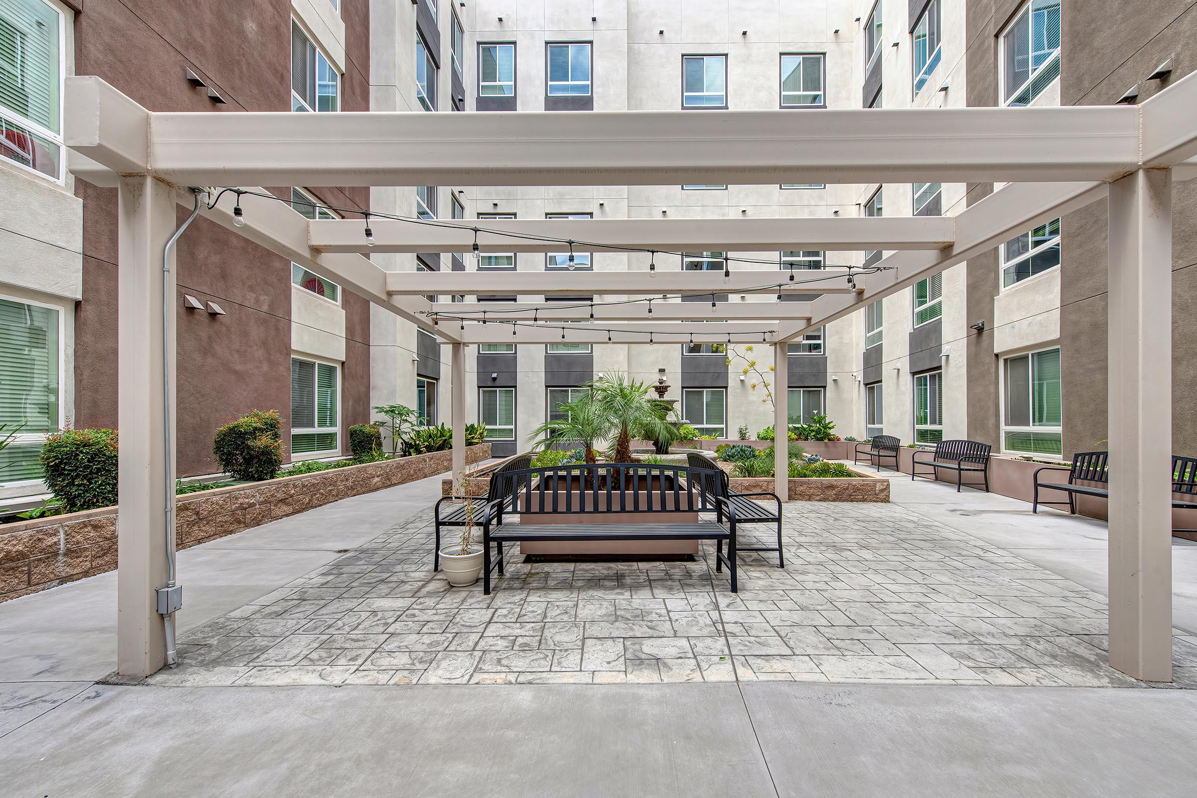 An outdoor courtyard featuring a pergola with string lights, a central bench, and landscaped areas with plants. The surrounding buildings are modern with multiple windows, creating a serene environment for relaxation or socializing.