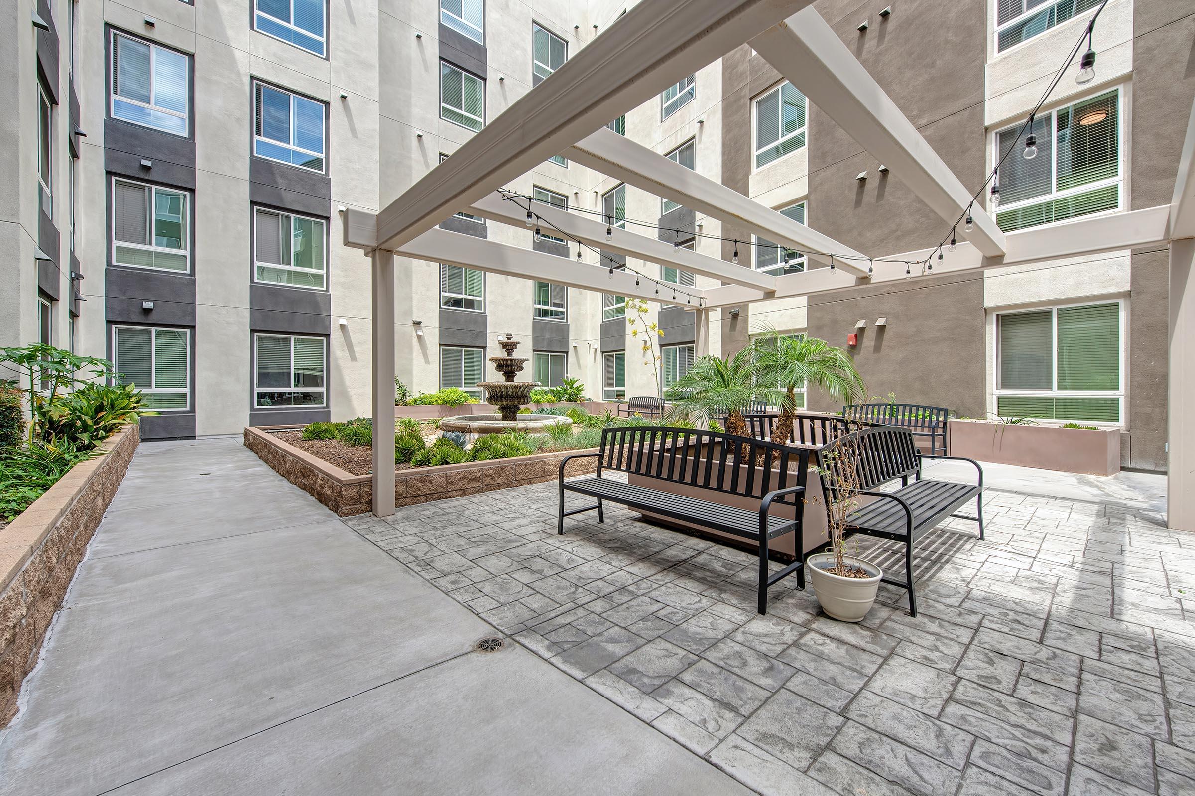 A landscaped courtyard featuring a fountain surrounded by seating areas. Modern buildings with large windows frame the space, which includes greenery, planters, and string lights overhead, creating a serene and inviting atmosphere.