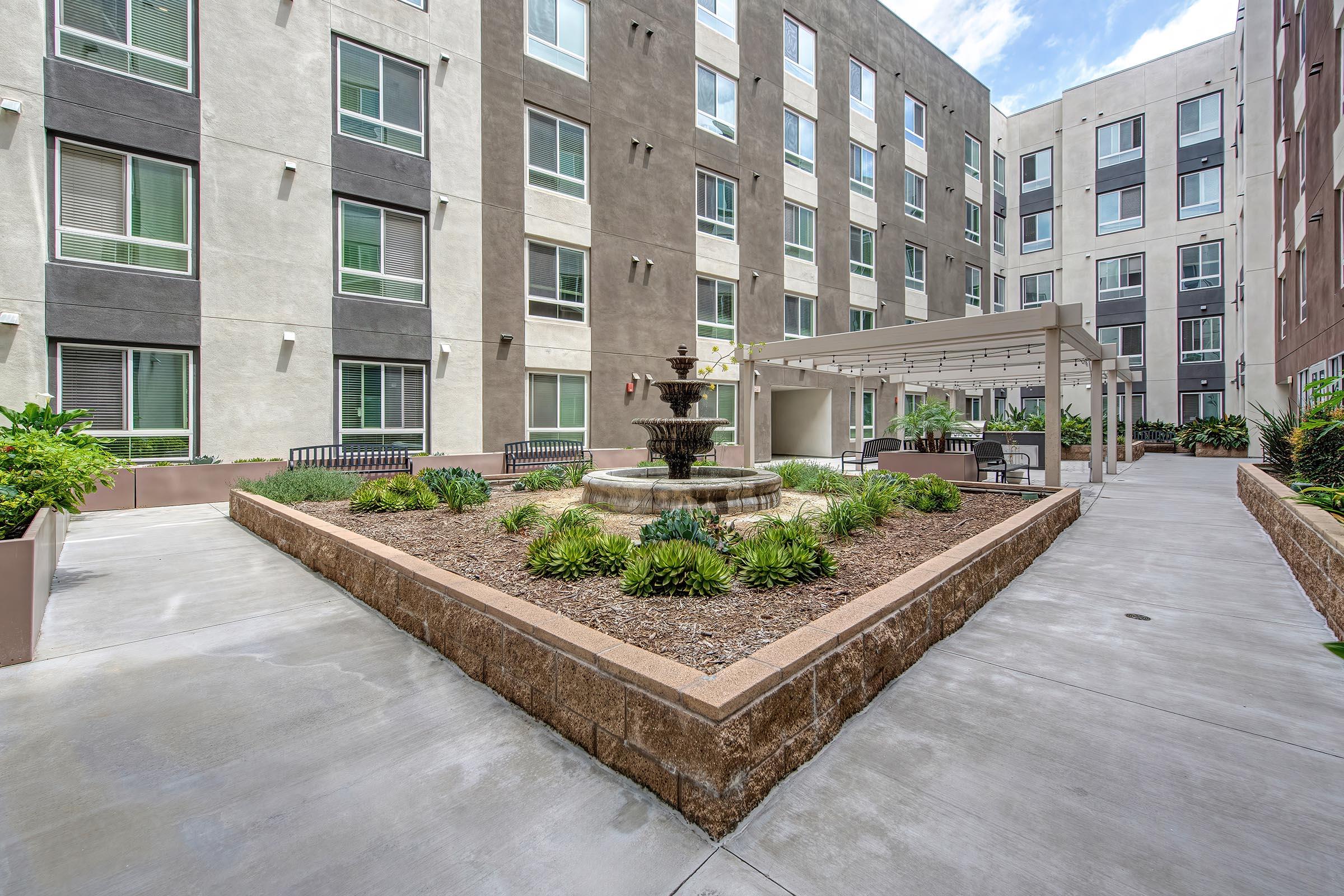 A modern courtyard featuring a central fountain surrounded by greenery and seating areas. The surrounding buildings have a mix of light and dark exterior colors, with large windows that let in natural light. The pathway is paved and well-maintained, creating an inviting outdoor space.