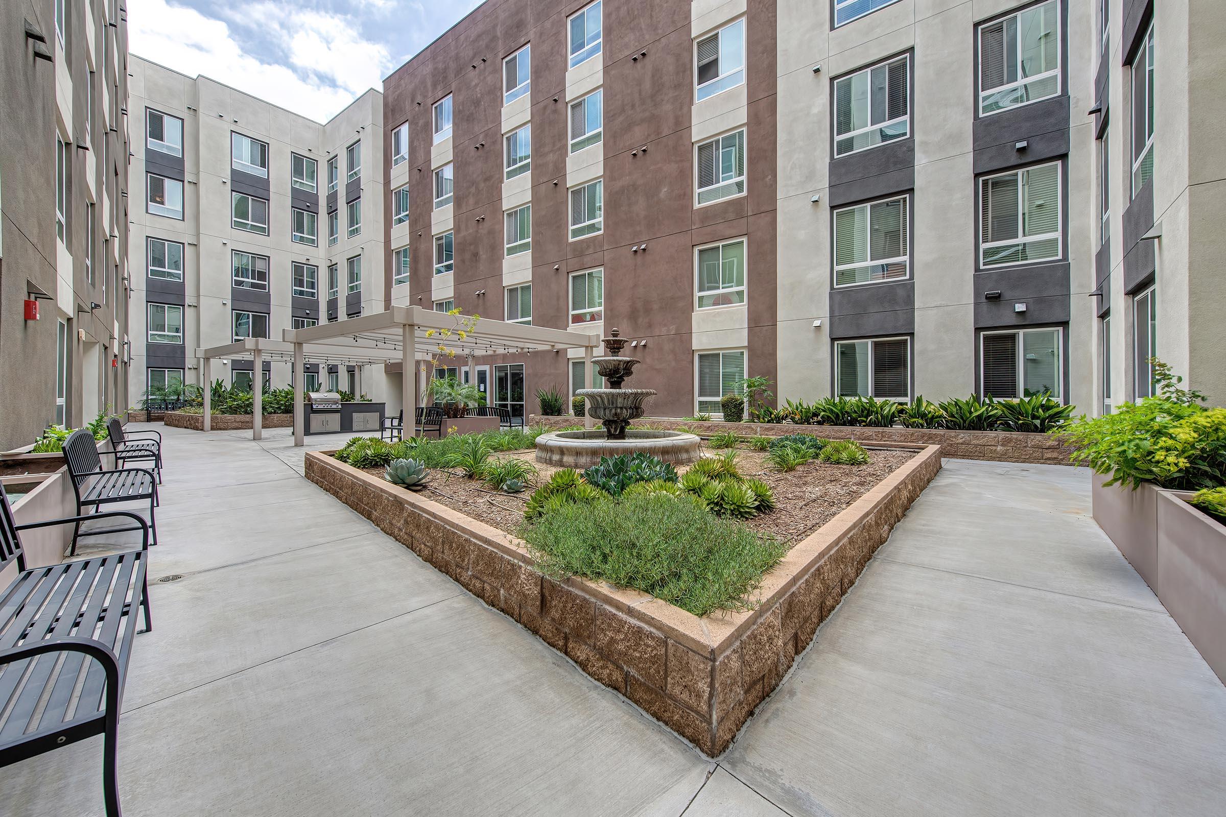 A landscaped courtyard featuring a central fountain surrounded by greenery and planters, with several benches lining the pathways. The backdrop consists of modern multi-story buildings with large windows, providing a bright and inviting atmosphere.