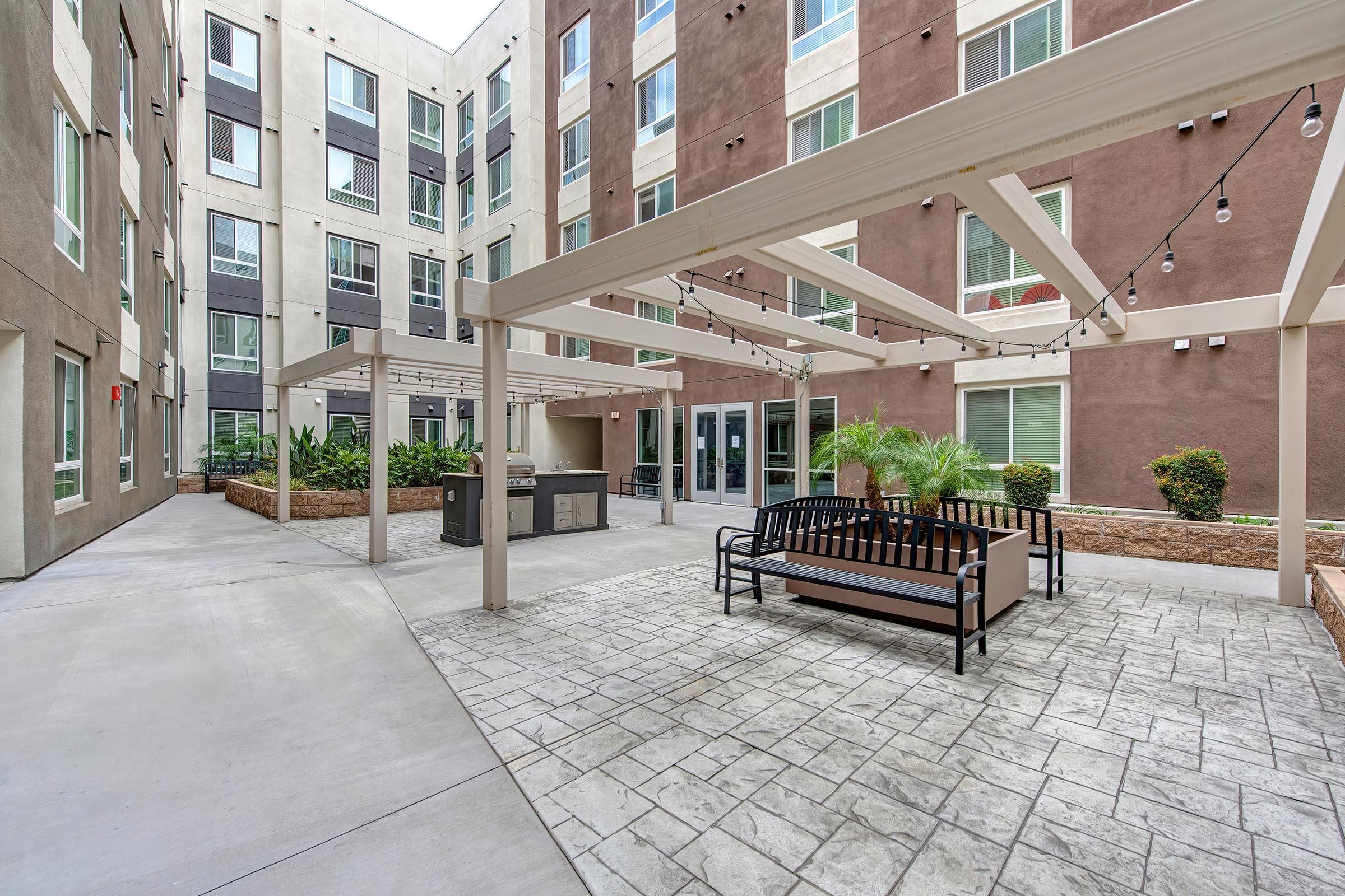 A modern outdoor courtyard featuring a paved area with benches, overhead string lights, and lush greenery. The space is surrounded by multi-story buildings with large windows, creating a welcoming atmosphere for relaxation and social gatherings.
