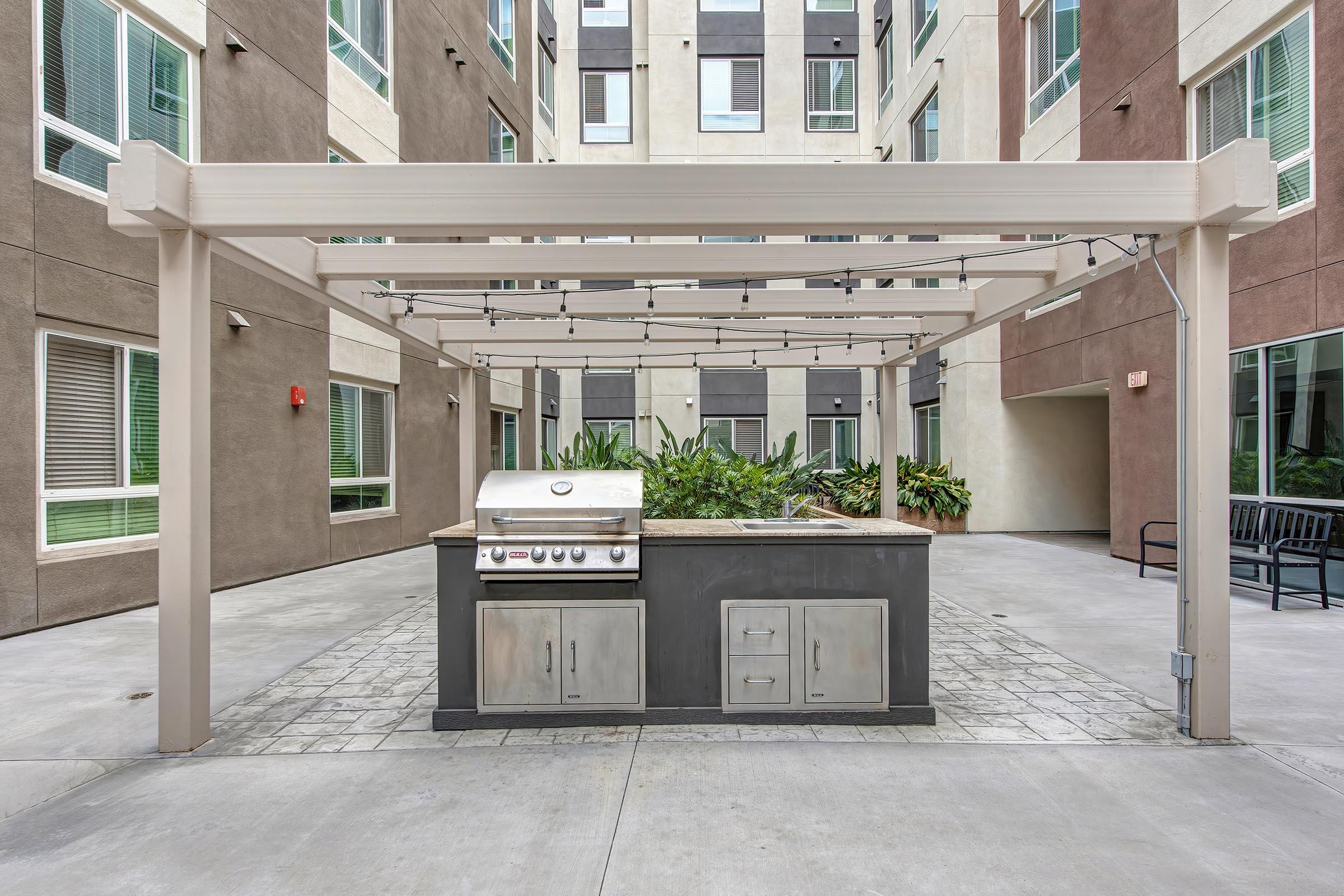 Outdoor barbecue area featuring a stainless steel grill and a counter space, situated under a pergola with string lights. Surrounding the grill are lush green plants and modern apartment buildings provide a backdrop, creating a welcoming communal space for cooking and gathering.