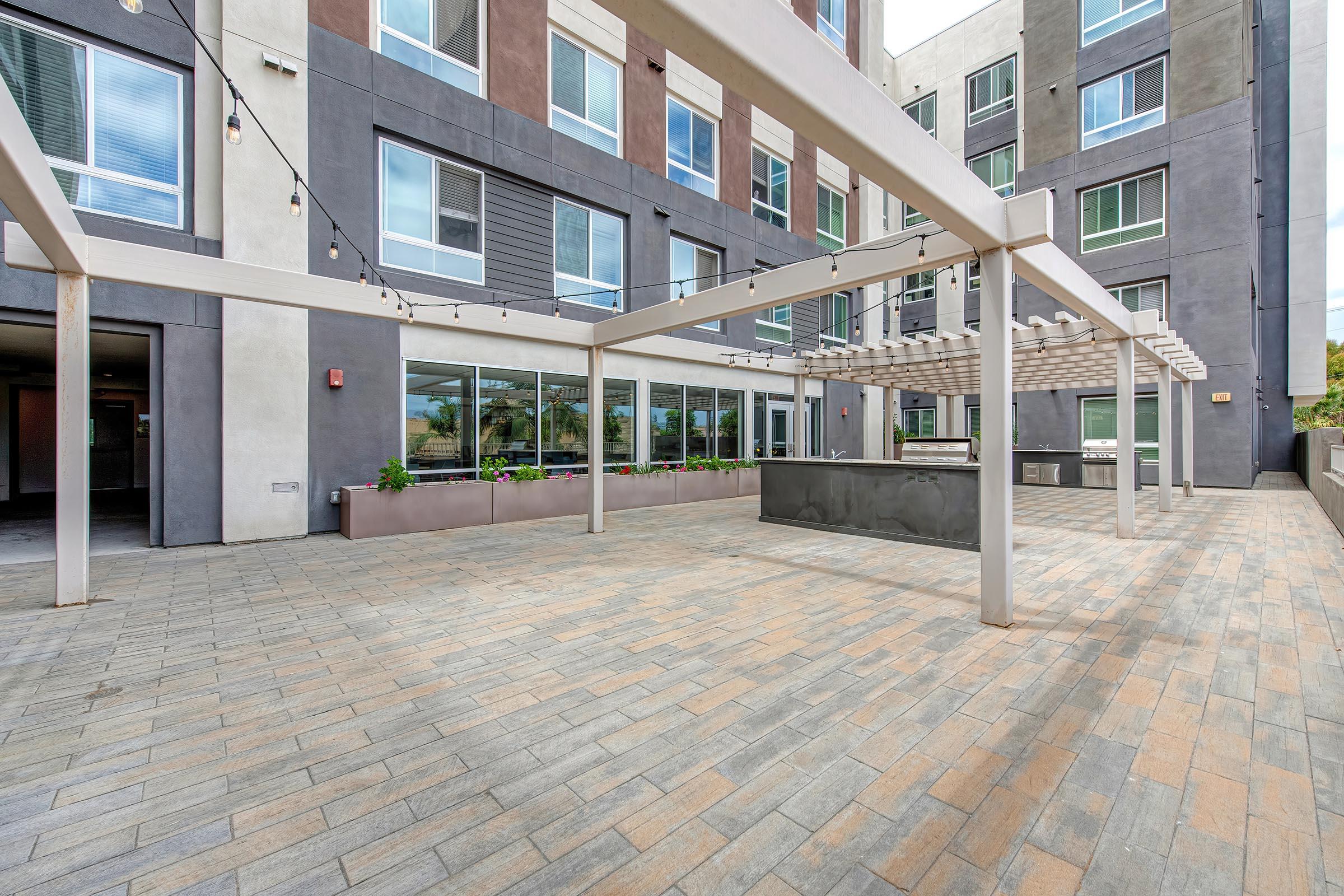 An outdoor patio area with modern design features, including light fixtures strung overhead and a large concrete structure with seating. The floor is paved with rectangular tiles, and the background showcases a contemporary building with large windows.