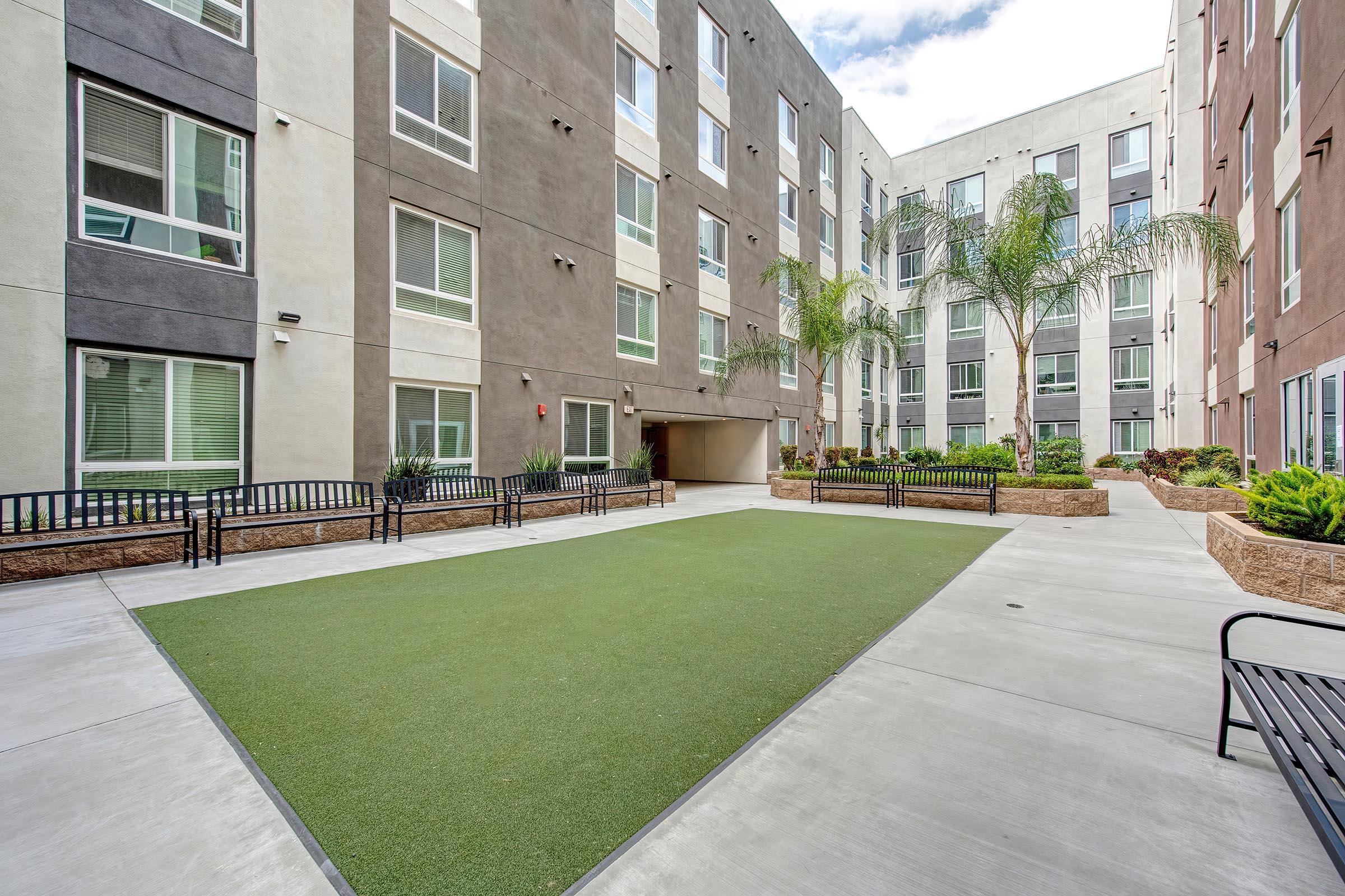 A well-maintained courtyard of an apartment complex featuring green artificial grass, bordered by benches and landscaped planters. Surrounding the courtyard are modern multi-story buildings with large windows, providing natural light to the area, and palm trees adding to the aesthetic appeal.