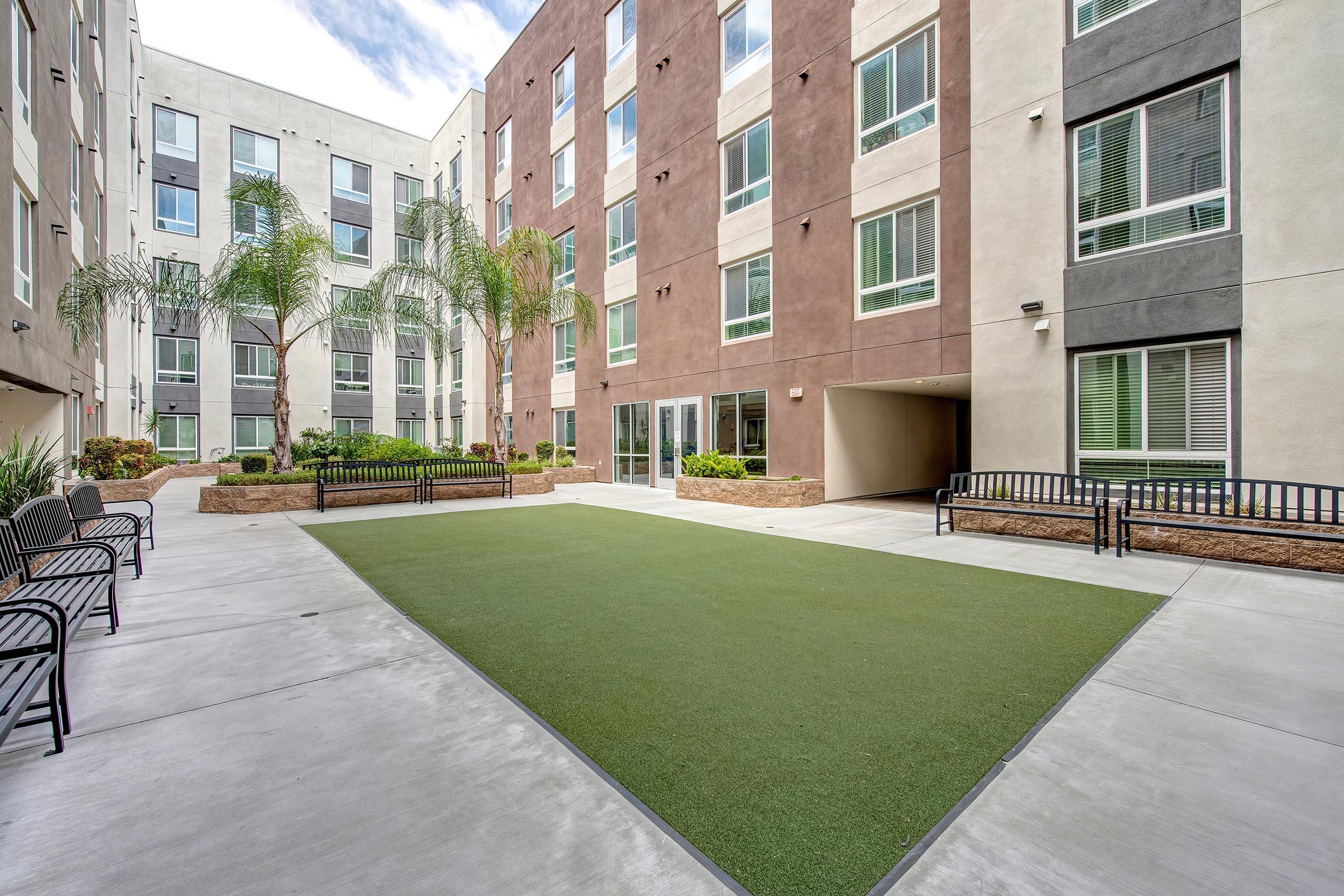 A landscaped courtyard featuring artificial grass, palm trees, and benches. The surrounding buildings have modern architecture with a mix of light and dark brown exteriors. Large windows reflect the sky, creating an inviting outdoor space.