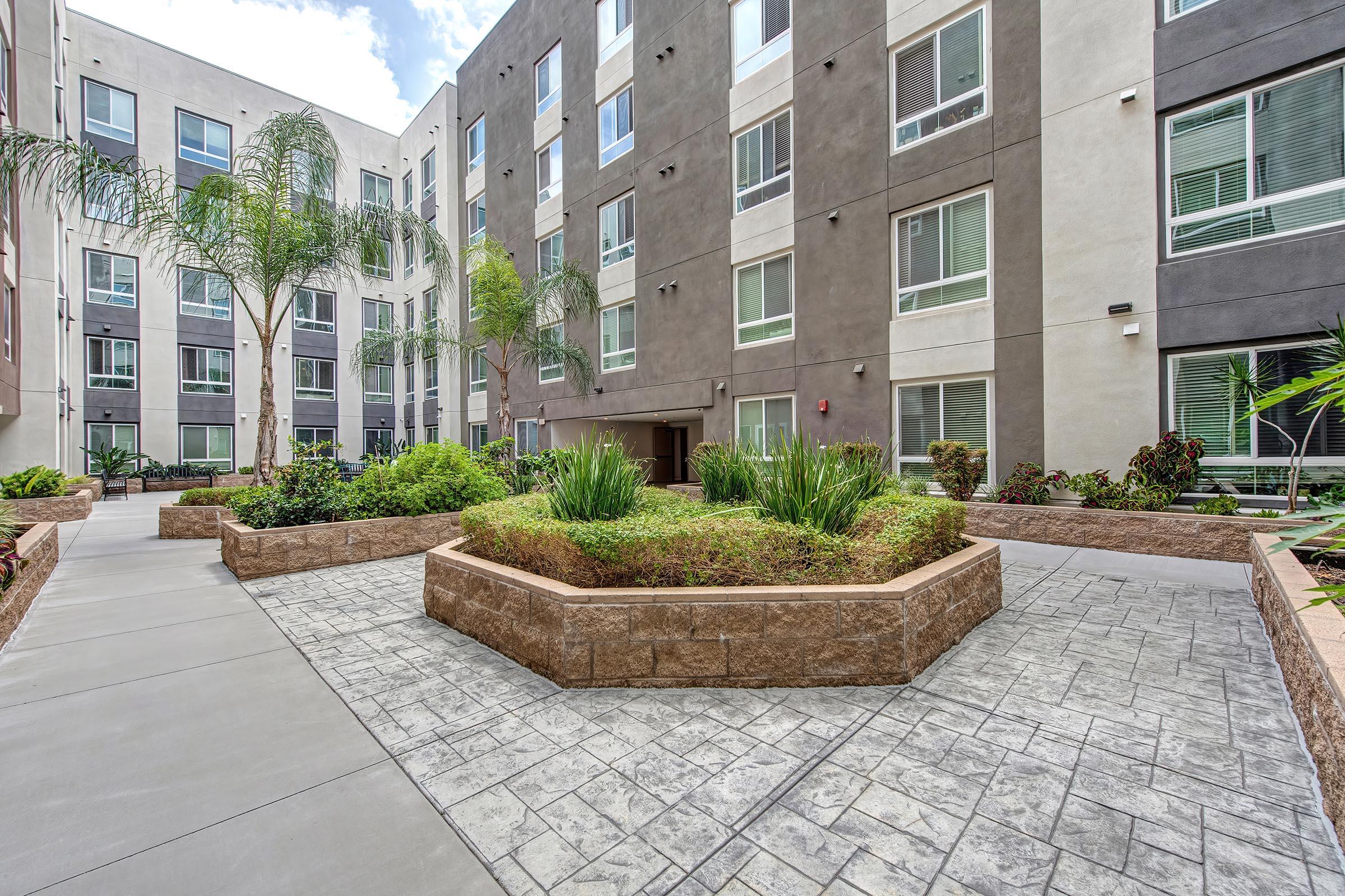 A landscaped courtyard area featuring a central planter with greenery, surrounded by modern multi-story buildings with large windows. The pathway is paved and leads through the garden, with palm trees and other plants enhancing the outdoor space.