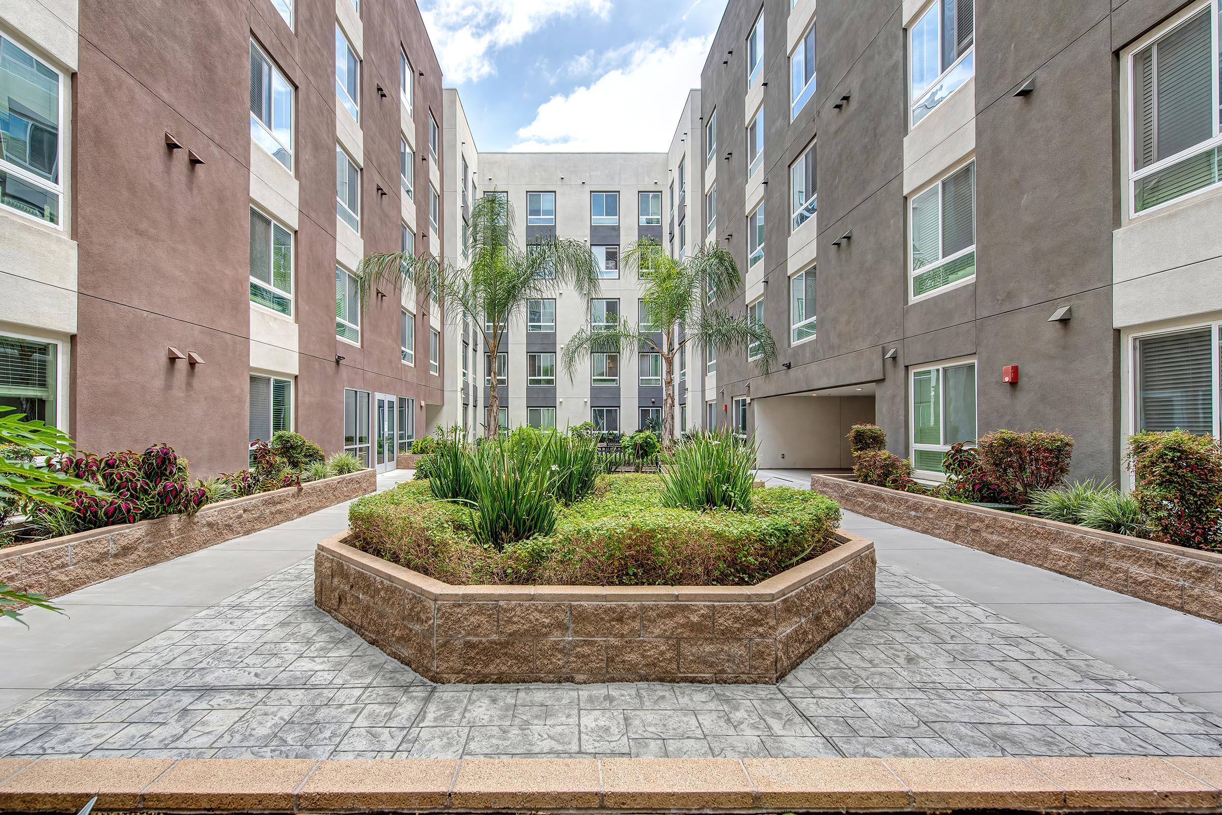 A landscaped courtyard featuring a central garden surrounded by modern apartment buildings. The area includes palm trees and decorative shrubs, with a paved walkway leading through the space. The sky is partly cloudy, enhancing the bright and inviting atmosphere of the environment.