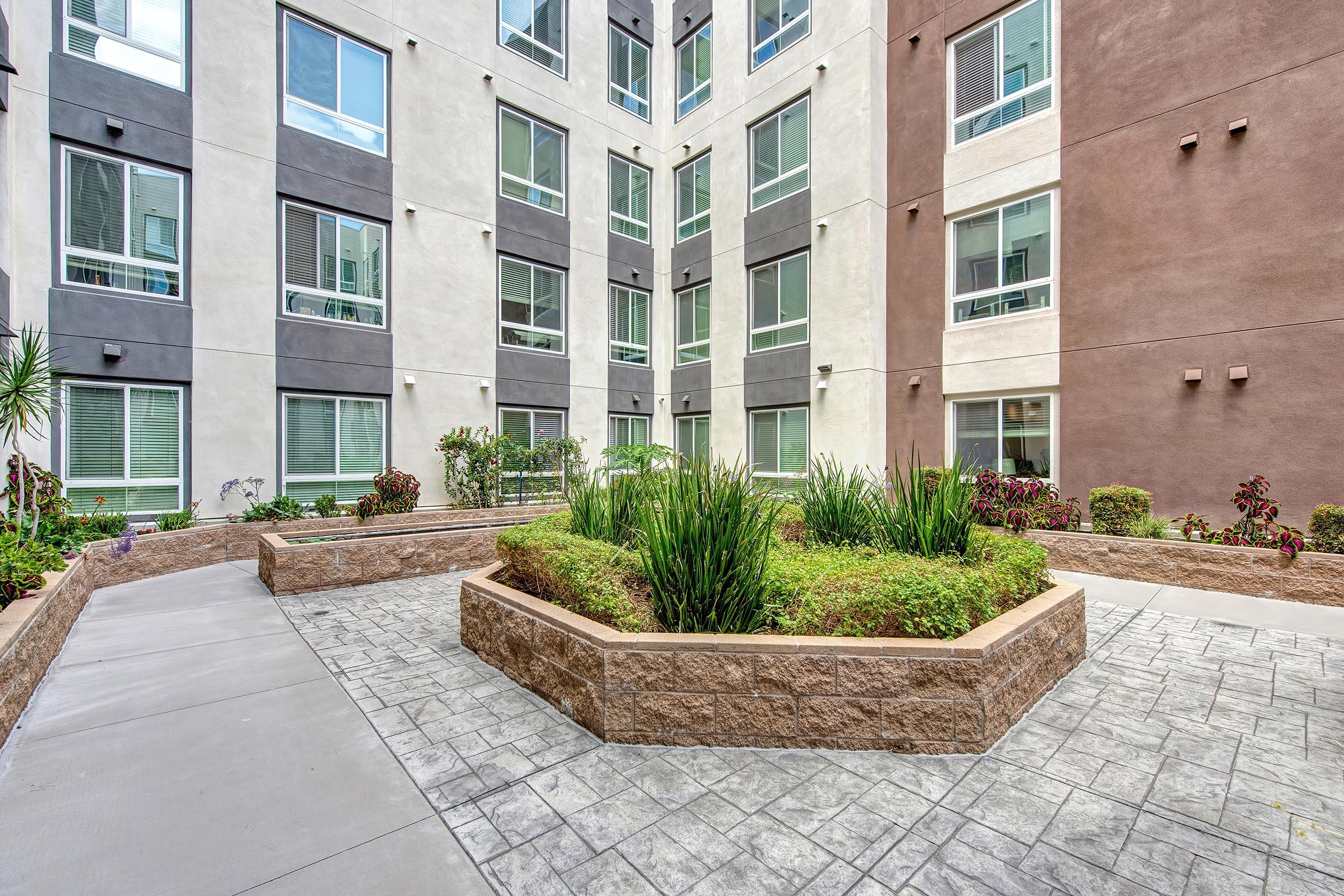 A modern courtyard surrounded by multi-story buildings, featuring neatly landscaped shrubs and plants in a raised garden bed. The ground is paved with decorative concrete, creating an inviting outdoor space with ample natural light and greenery.