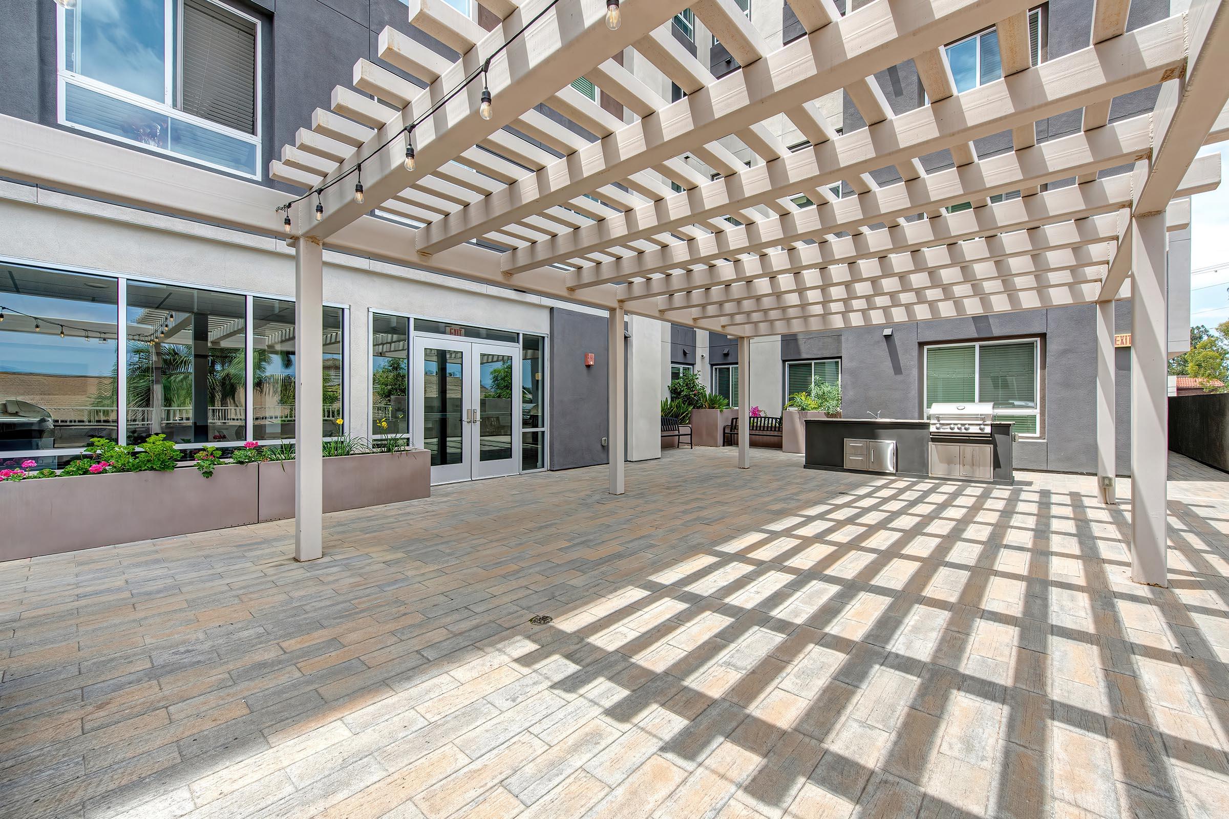 A spacious outdoor patio area featuring a wooden pergola, landscaped flower beds, and a built-in barbecue grill. The space is well-lit with shadows created by the pergola slats, providing a cozy atmosphere for gatherings. Large sliding glass doors are visible in the background, leading indoors.