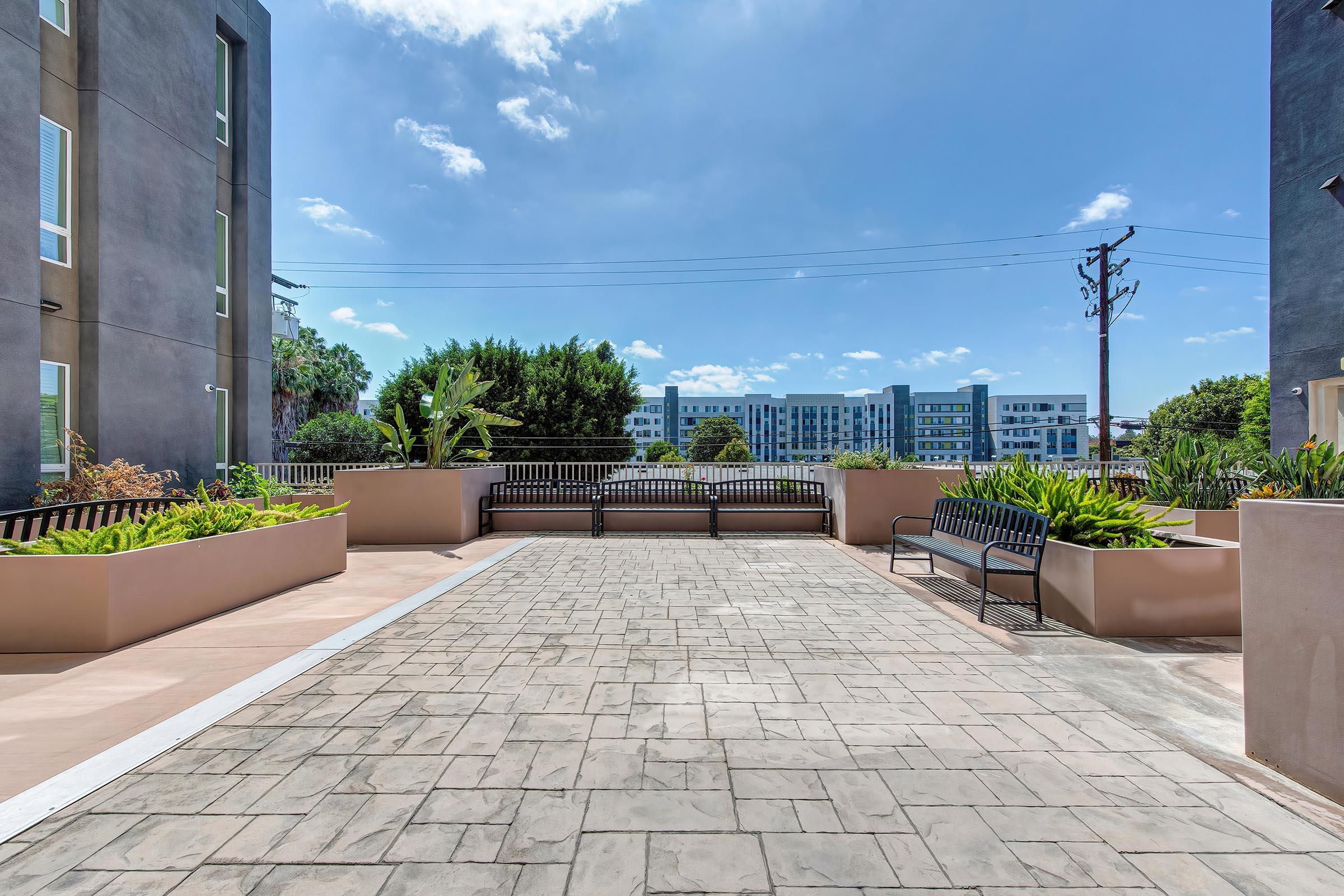 A modern outdoor patio area with concrete paving, bordered by lush green plants and low walls. Two benches are placed along the patio, with a clear blue sky overhead. In the background, contemporary buildings are visible, and there are power lines running across the scene.