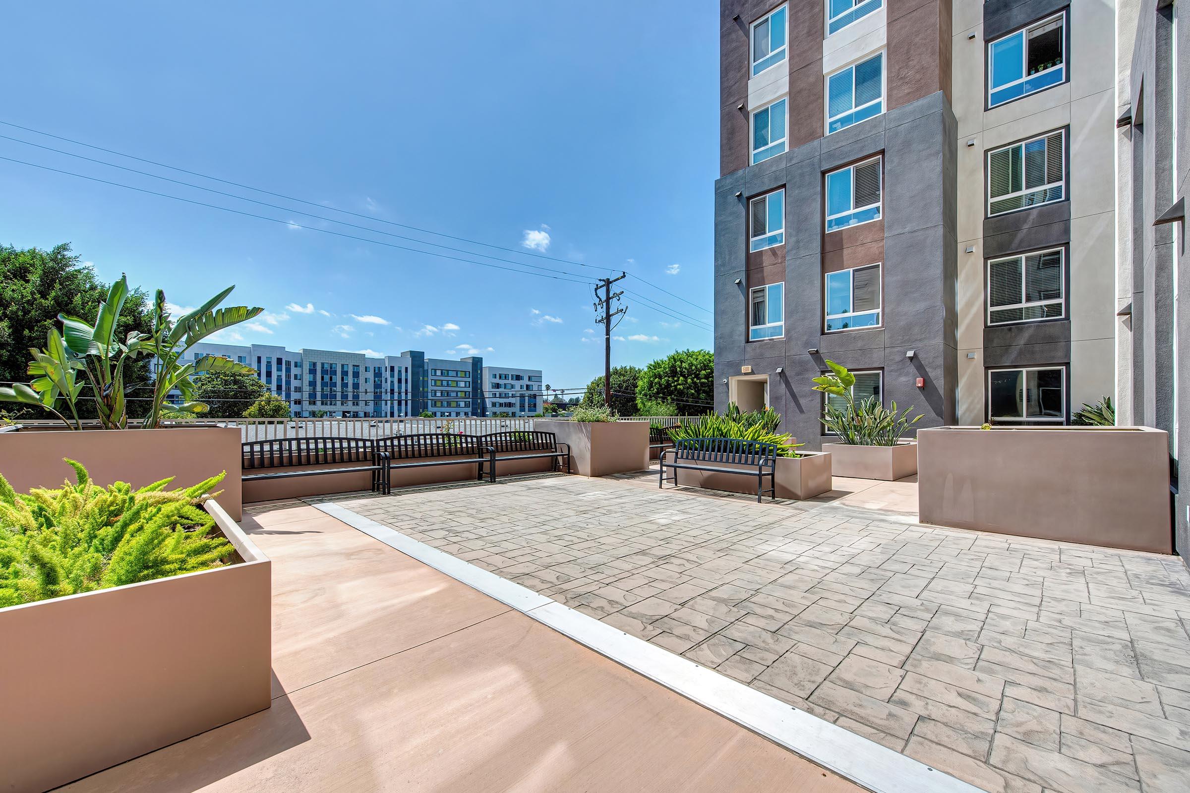A spacious outdoor patio area featuring modern landscaping with potted plants, benches, and a paved surface. The background showcases a clear blue sky and contemporary apartment buildings, creating a bright and inviting atmosphere. Ideal for relaxation and outdoor gatherings.