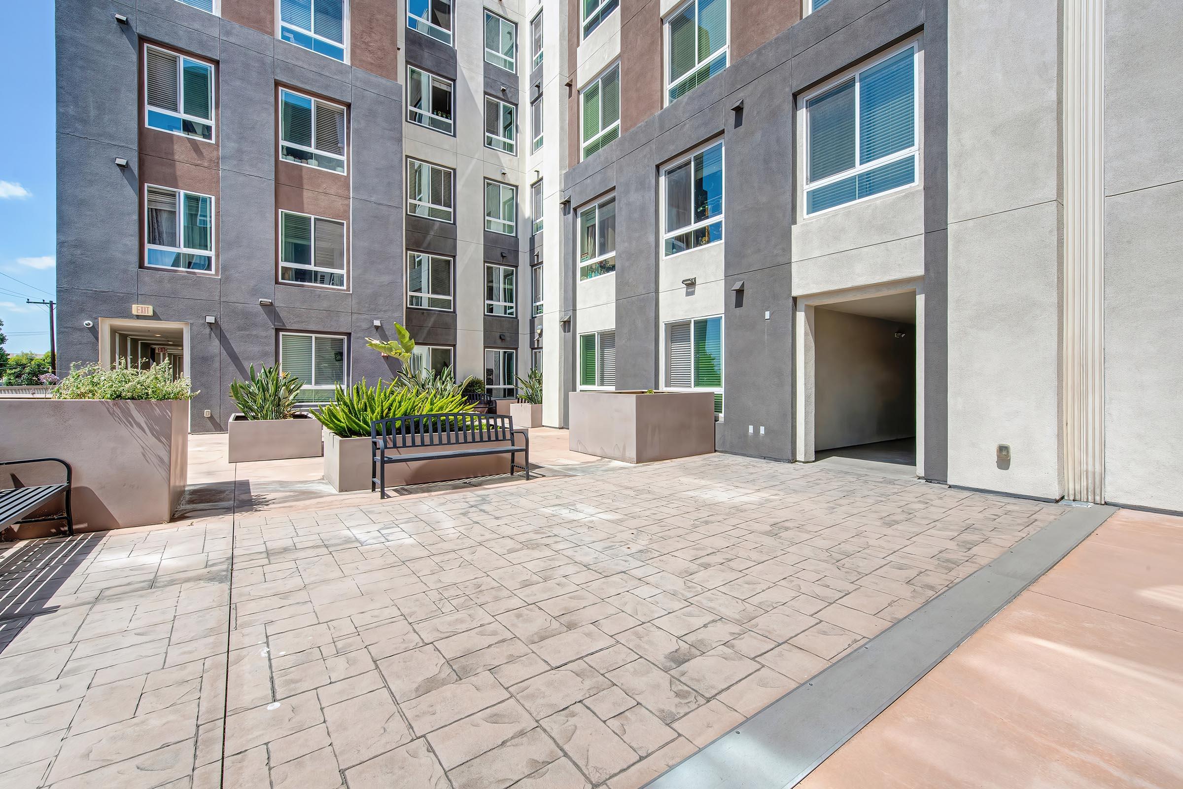 A modern apartment complex courtyard featuring a stamped concrete patio, planters with greenery, and a bench for seating. The building has a mix of brown and gray facade with large windows, letting in natural light. An entrance to the building is visible on the right side of the image.