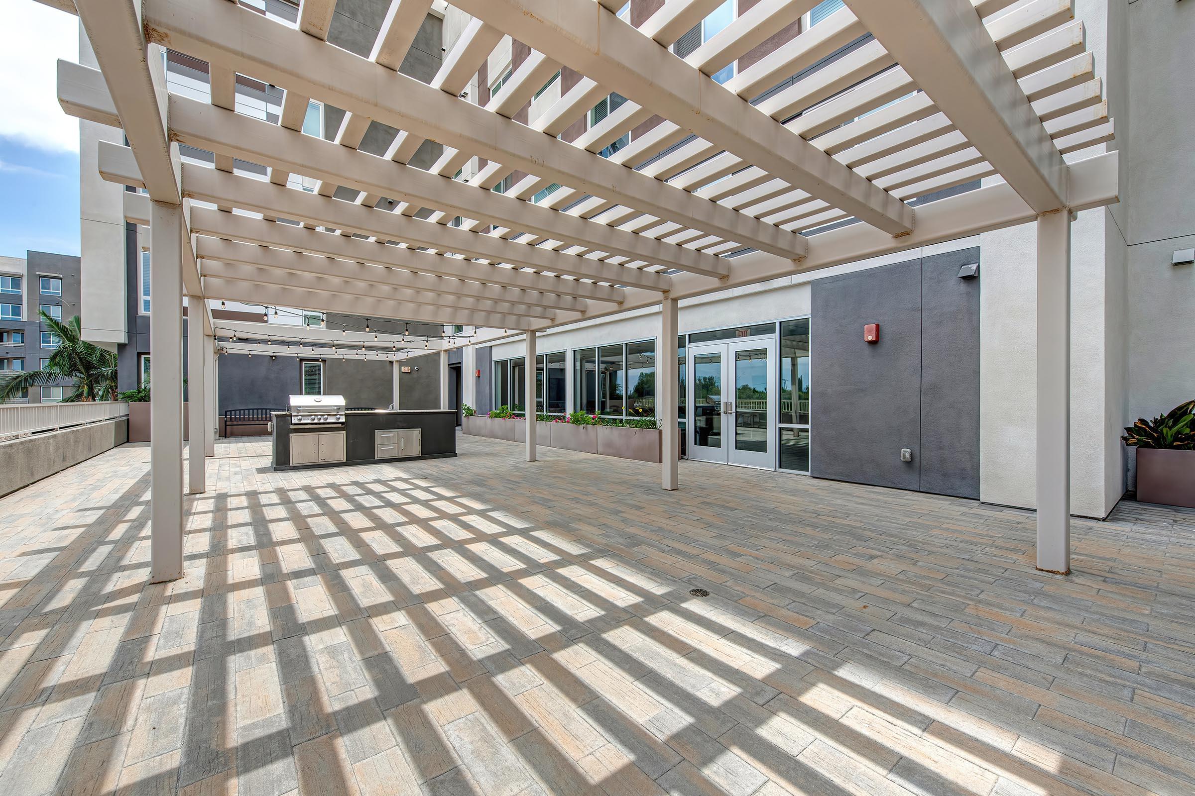 A spacious outdoor patio area with a pergola casting shadows on the tiled floor. There are outdoor dining facilities including a grill. Surrounding the area are modern buildings, and the space is well-lit with greenery in planters. Large glass doors lead into the building.