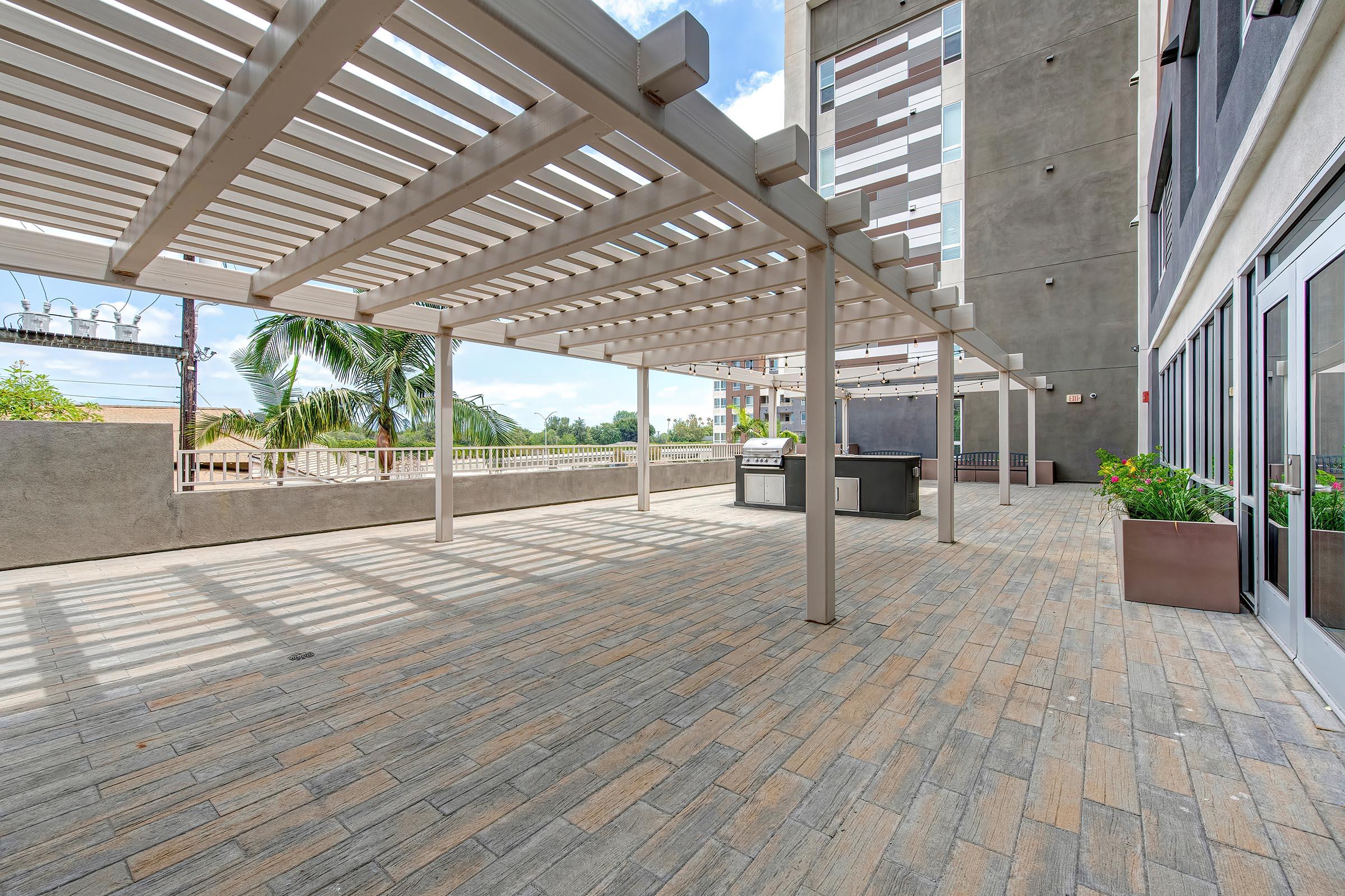 A spacious outdoor patio area featuring a light-colored pergola with horizontal slats, surrounded by potted plants. There is a grill station against a wall, and the floor is paved with large rectangular tiles. The background includes a view of palm trees and a modern building facade.
