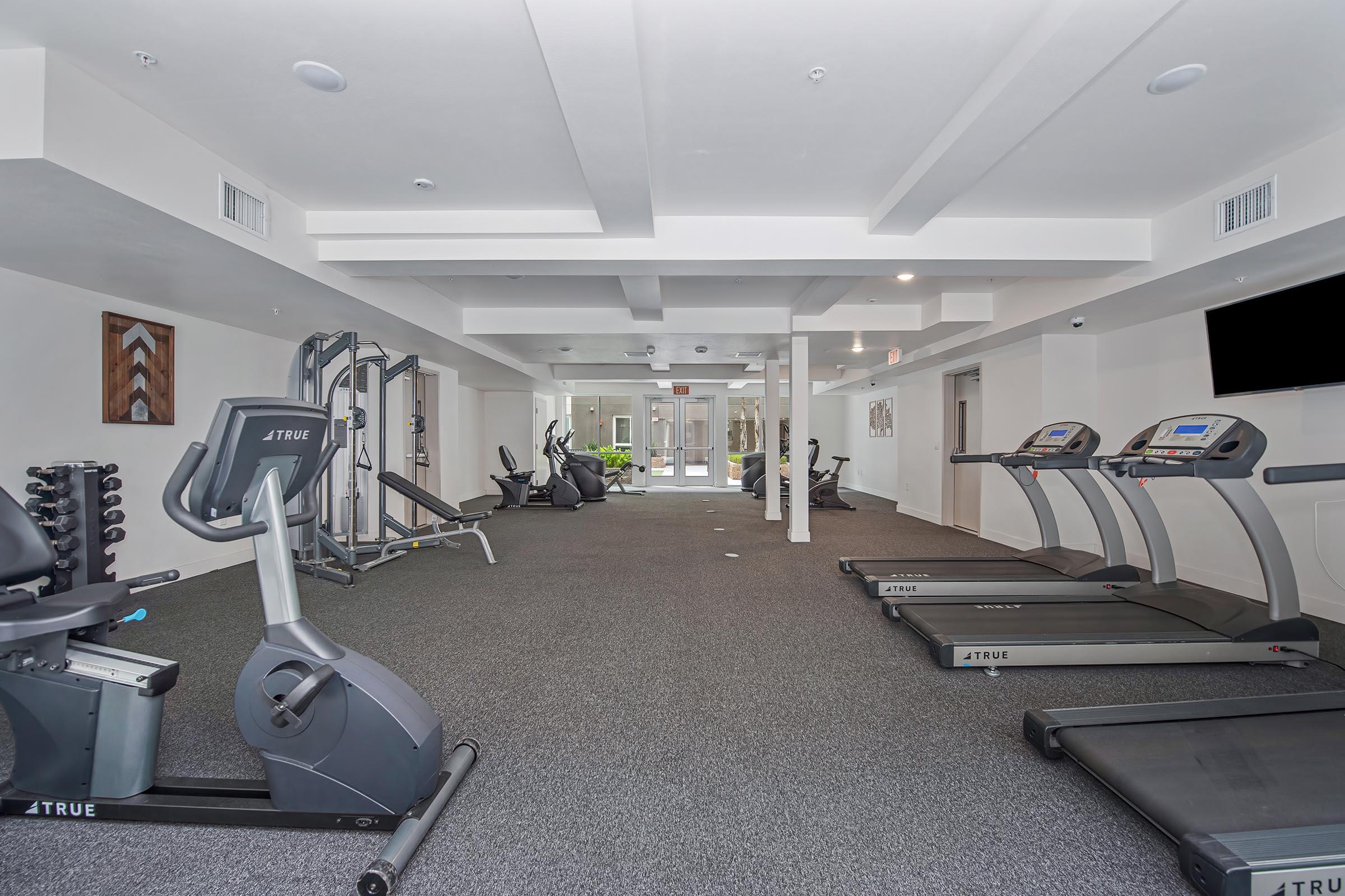 A modern gym interior featuring various exercise equipment including treadmills, stationary bikes, and weight machines. The space has carpeted flooring, bright lighting, and large windows allowing natural light to enter. The atmosphere is clean and inviting for workout activities.