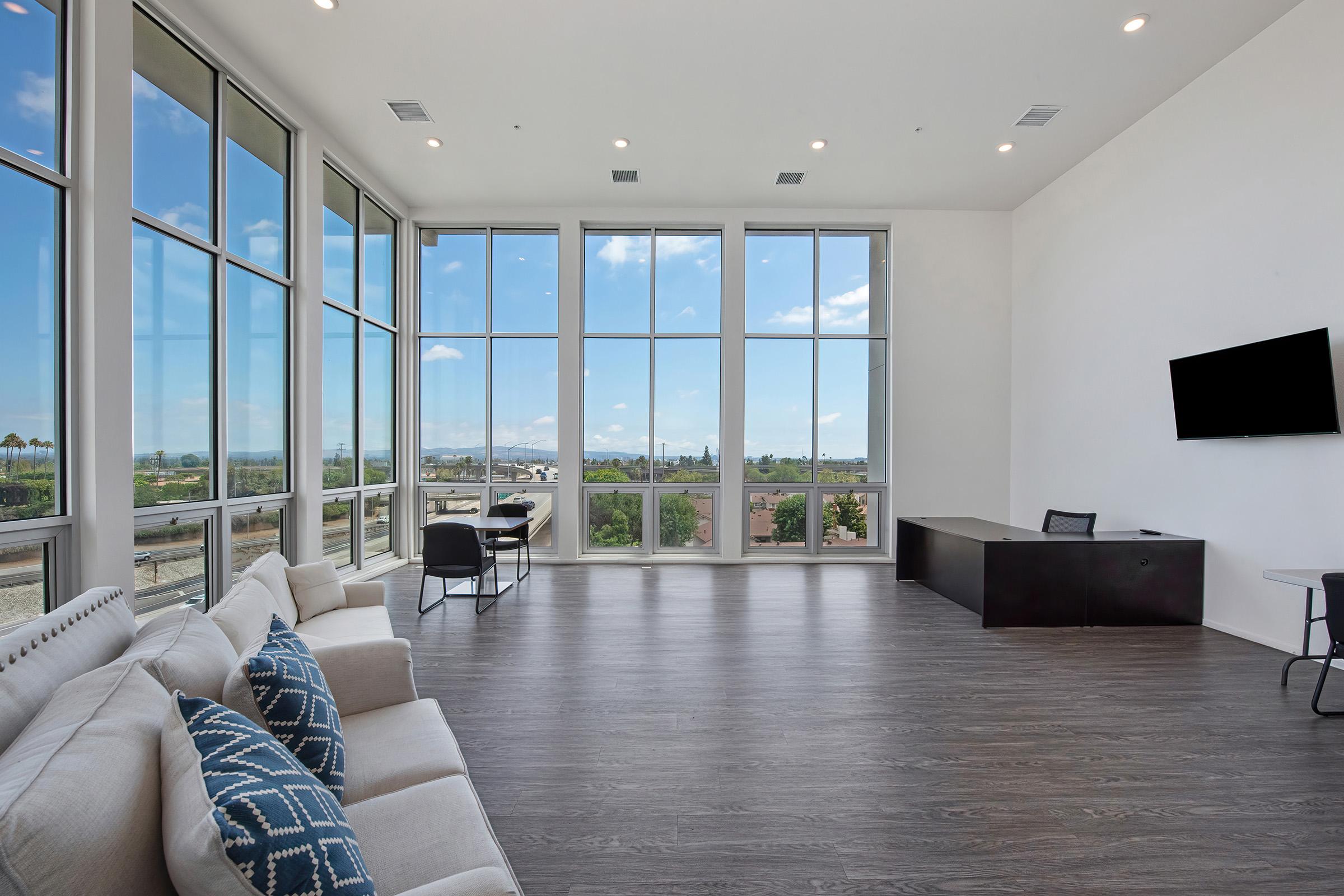 A spacious, modern interior featuring large windows that let in natural light. The room includes a light-colored sofa with decorative pillows, a black desk and chair, and a mounted flat-screen TV. A view of the outdoors is visible through the windows, showcasing a clear blue sky.