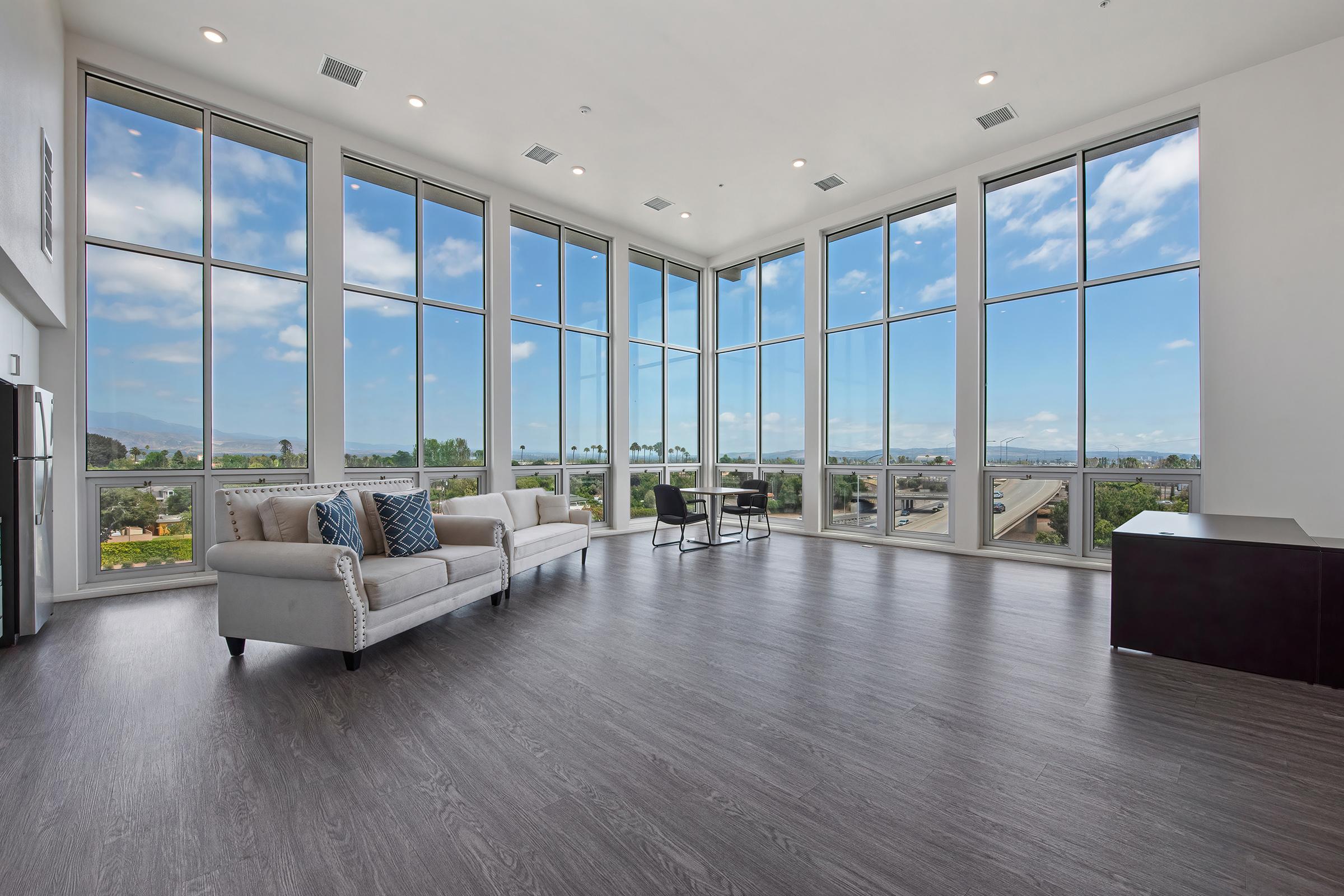 A spacious, modern living room with large floor-to-ceiling windows showcasing a scenic view. The room features light wood flooring, a beige sofa with decorative pillows, a black coffee table, and a minimalistic design, creating a bright and airy atmosphere.