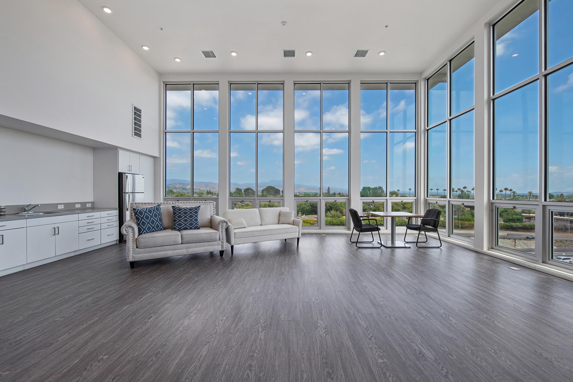 A modern, spacious living area with large windows providing a panoramic view of the outdoors. The room features a light-colored sofa, a black chair, and a sleek kitchenette. Brightly lit by natural light, the flooring is dark wood, creating a contemporary and inviting atmosphere.