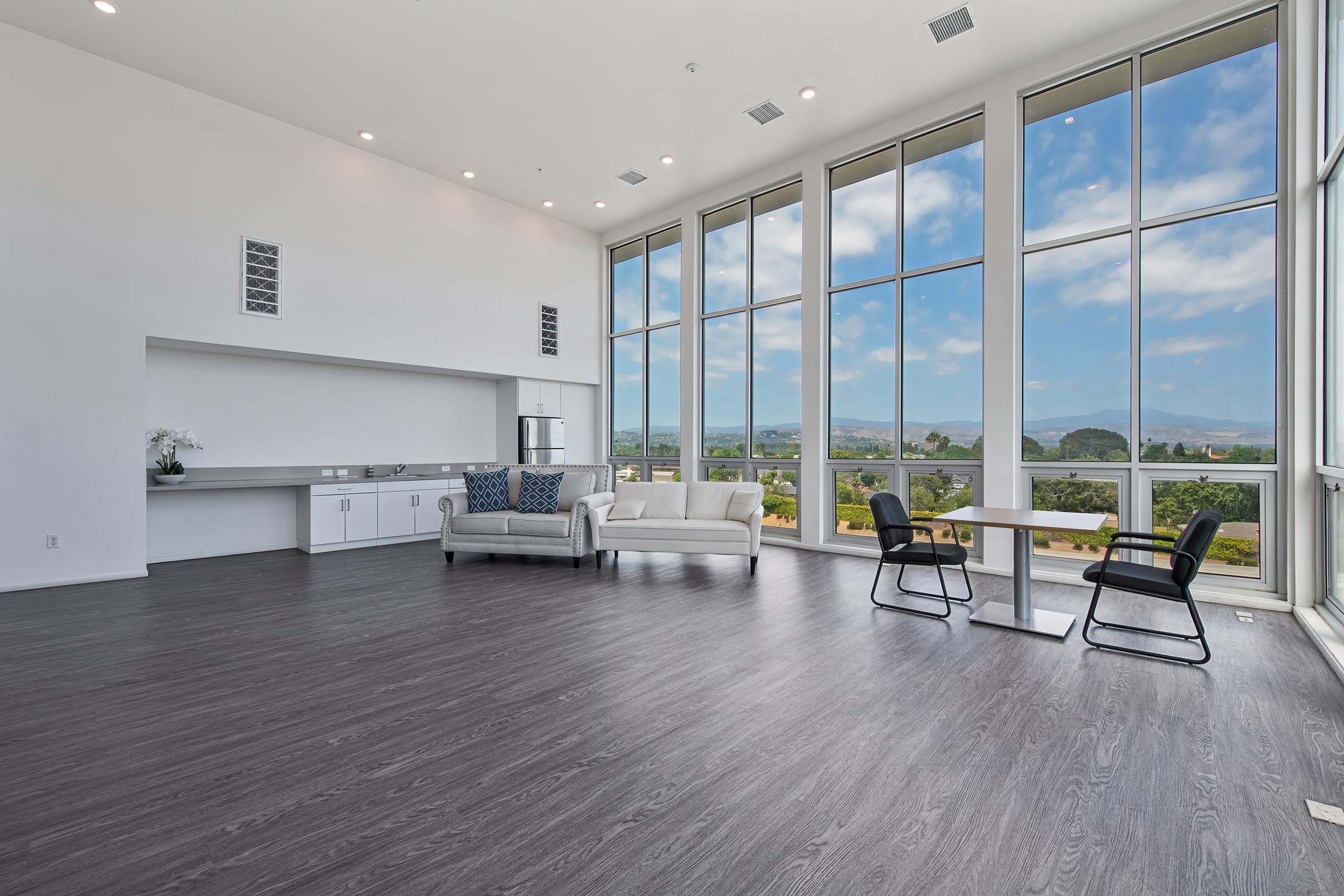 A spacious, modern interior with large floor-to-ceiling windows offering a scenic view of rolling hills and a blue sky. The room features light-colored walls and hardwood flooring, a sofa with decorative pillows, a dining table with chairs, and a minimalist kitchen area. Natural light floods the space, creating an inviting atmosphere.