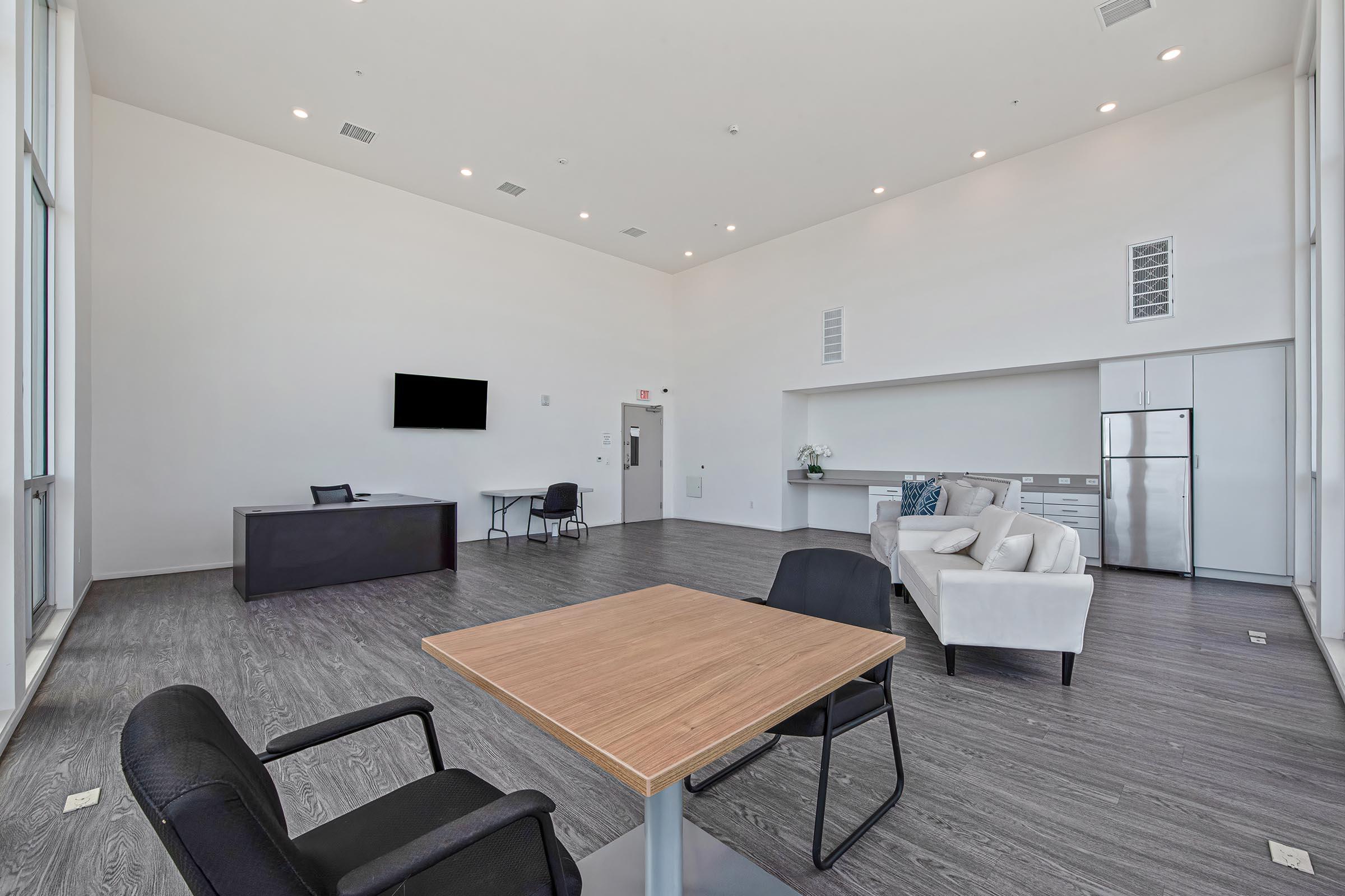 Spacious modern room with high ceilings, featuring a large window, a rectangular table with chairs, a light-colored sofa, and a kitchenette area. A wall-mounted TV is present, along with a desk and a refrigerator. The flooring is dark wood, creating a contemporary atmosphere.