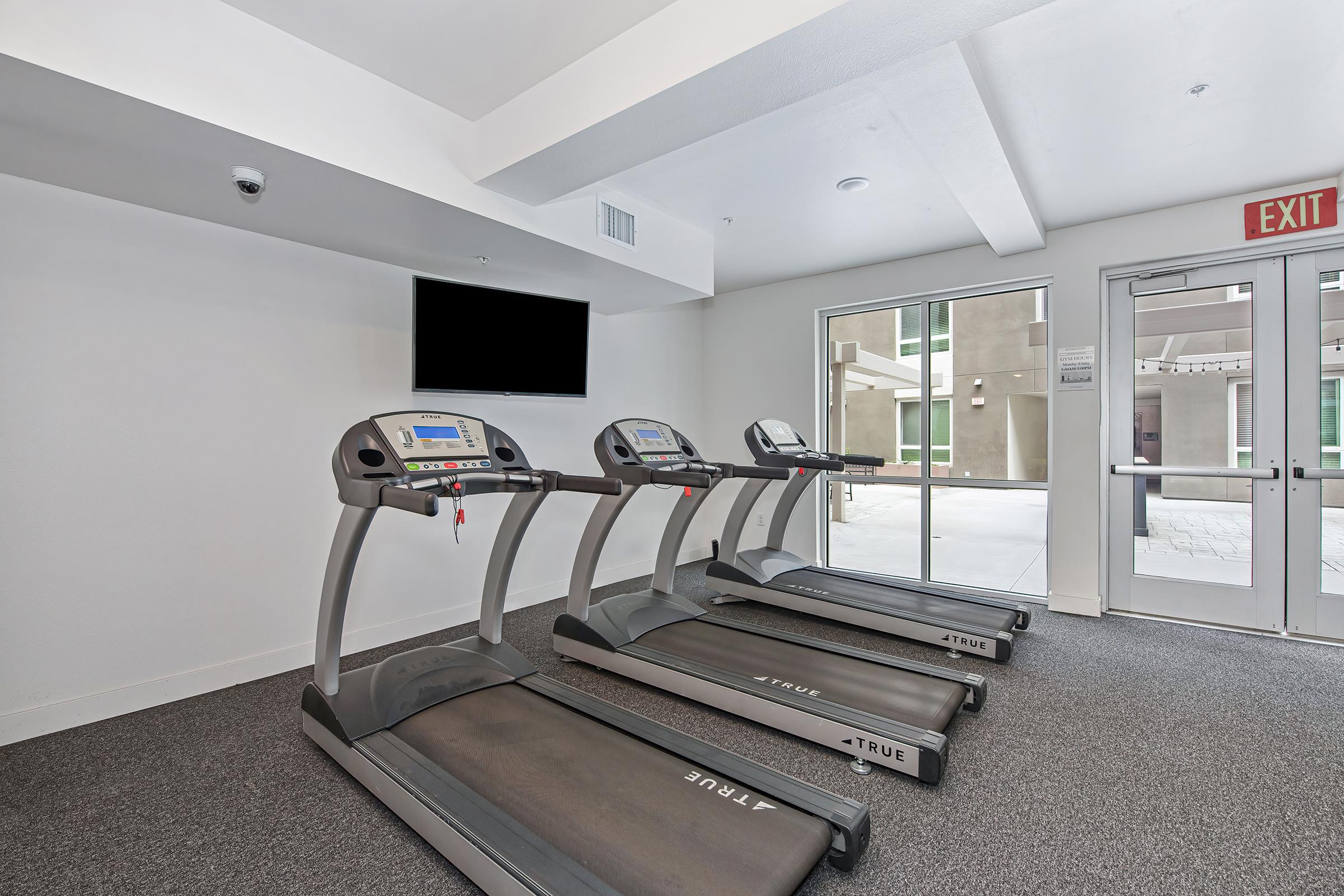 A fitness area featuring three treadmills lined up in front of a blank television screen. Large windows provide natural light, and the floor is carpeted. An exit door is visible on the right side. The overall atmosphere is bright and modern, ideal for exercise and fitness activities.