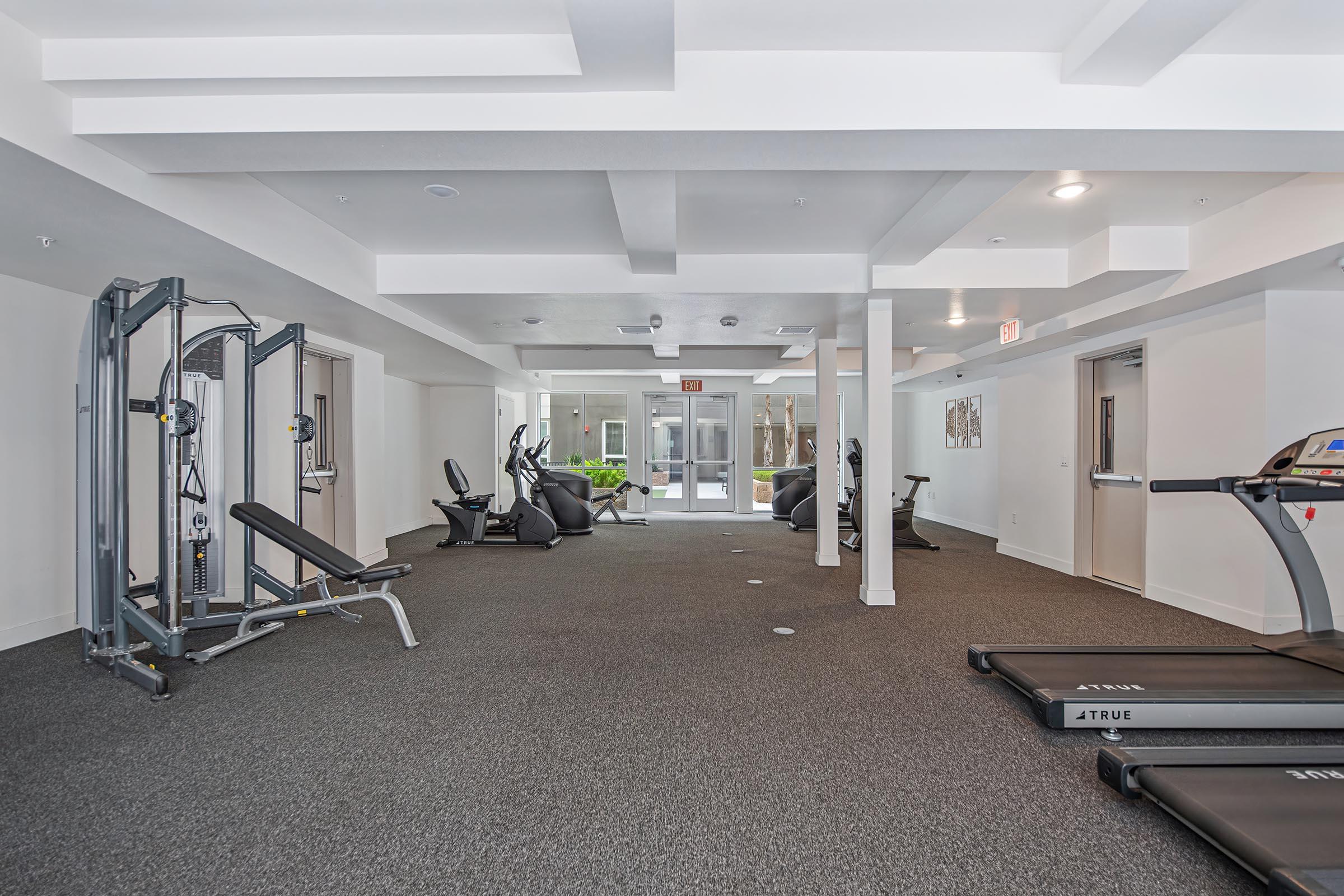 A spacious gym area featuring various exercise equipment, including treadmills, a weight machine, and stationary bikes. The floor is carpeted, and large windows allow natural light to enter, providing a bright and inviting atmosphere. The gym is well-maintained and modern.