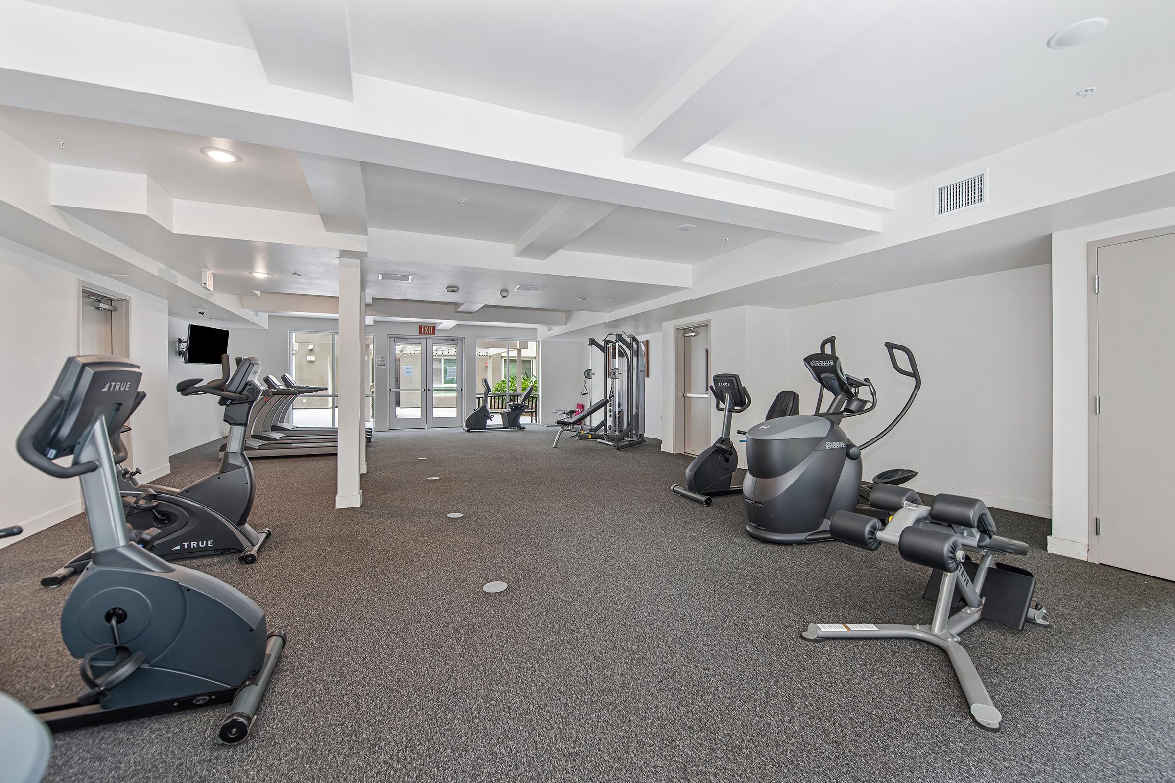 A modern indoor gym featuring various exercise equipment, including stationary bikes, treadmills, and weight machines. The space is well-lit with large windows, showcasing a clean and organized environment, ideal for fitness activities.
