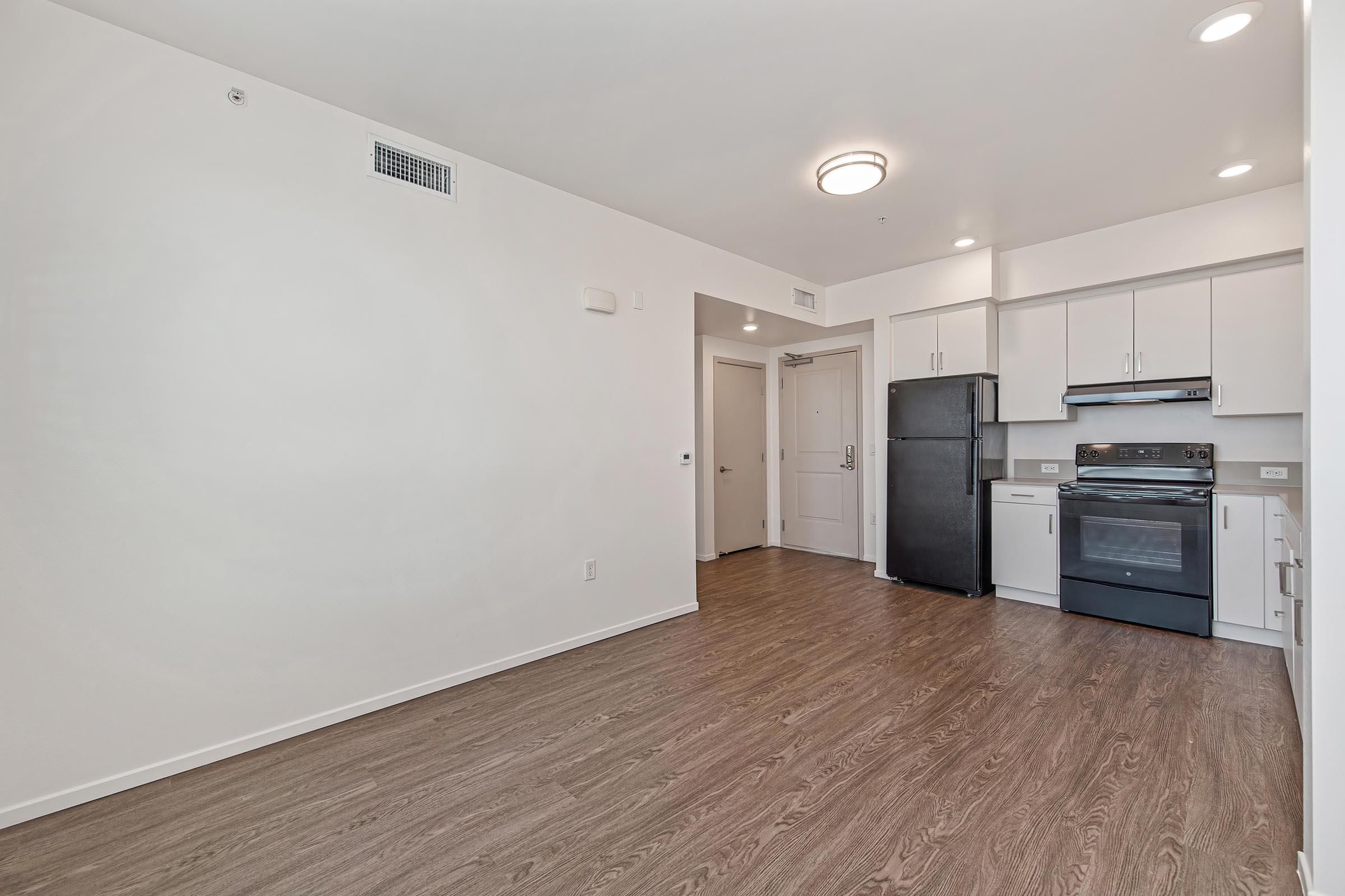 A modern studio apartment featuring an open layout with hardwood-style flooring. The kitchen includes a black refrigerator, an oven, and white cabinetry. There is a doorway leading to a bathroom and an entryway visible on the left side of the image. The walls are painted in a neutral color, and there is overhead lighting.