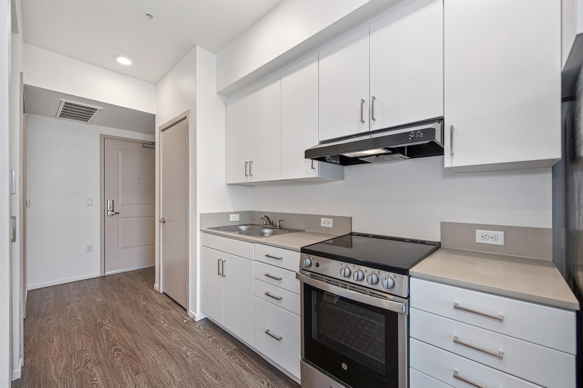 Modern kitchen featuring white cabinetry, stainless steel appliances, including a gas stove with an overhead microwave. The counter is a light color, and the flooring is wood-like. A door leads to a hallway, giving a view of the entryway. Bright and spacious environment with ample storage.