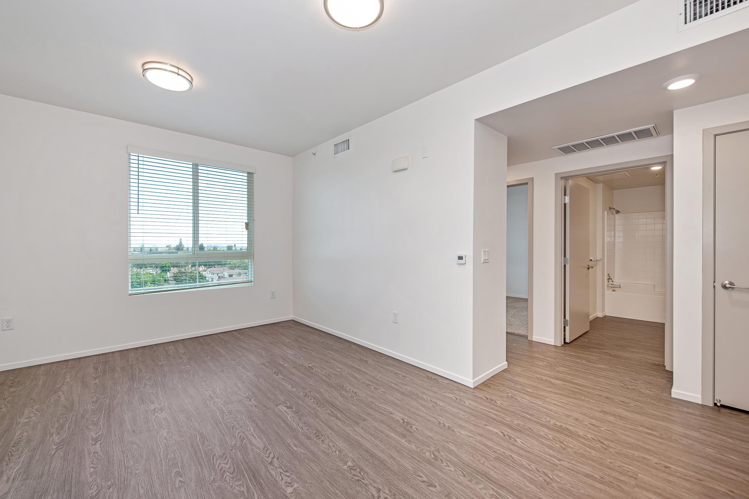 A modern, empty apartment interior featuring light-colored walls, hardwood flooring, and large windows allowing natural light. One corner shows a doorway leading to a small bathroom area, while another door opens into a hallway. The overall space is bright and minimalist, suitable for new furnishings.