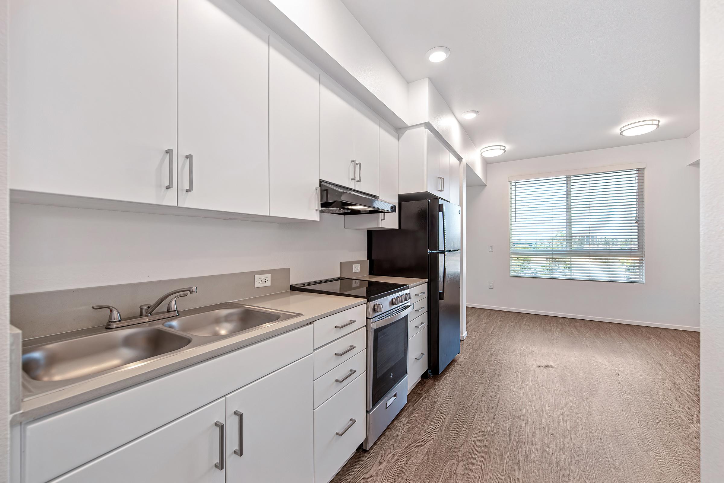 A modern kitchen featuring white cabinetry, stainless steel appliances including a stove and refrigerator, dual sinks, and a window with blinds allowing natural light. The flooring is a light wood style, creating a bright and airy atmosphere.