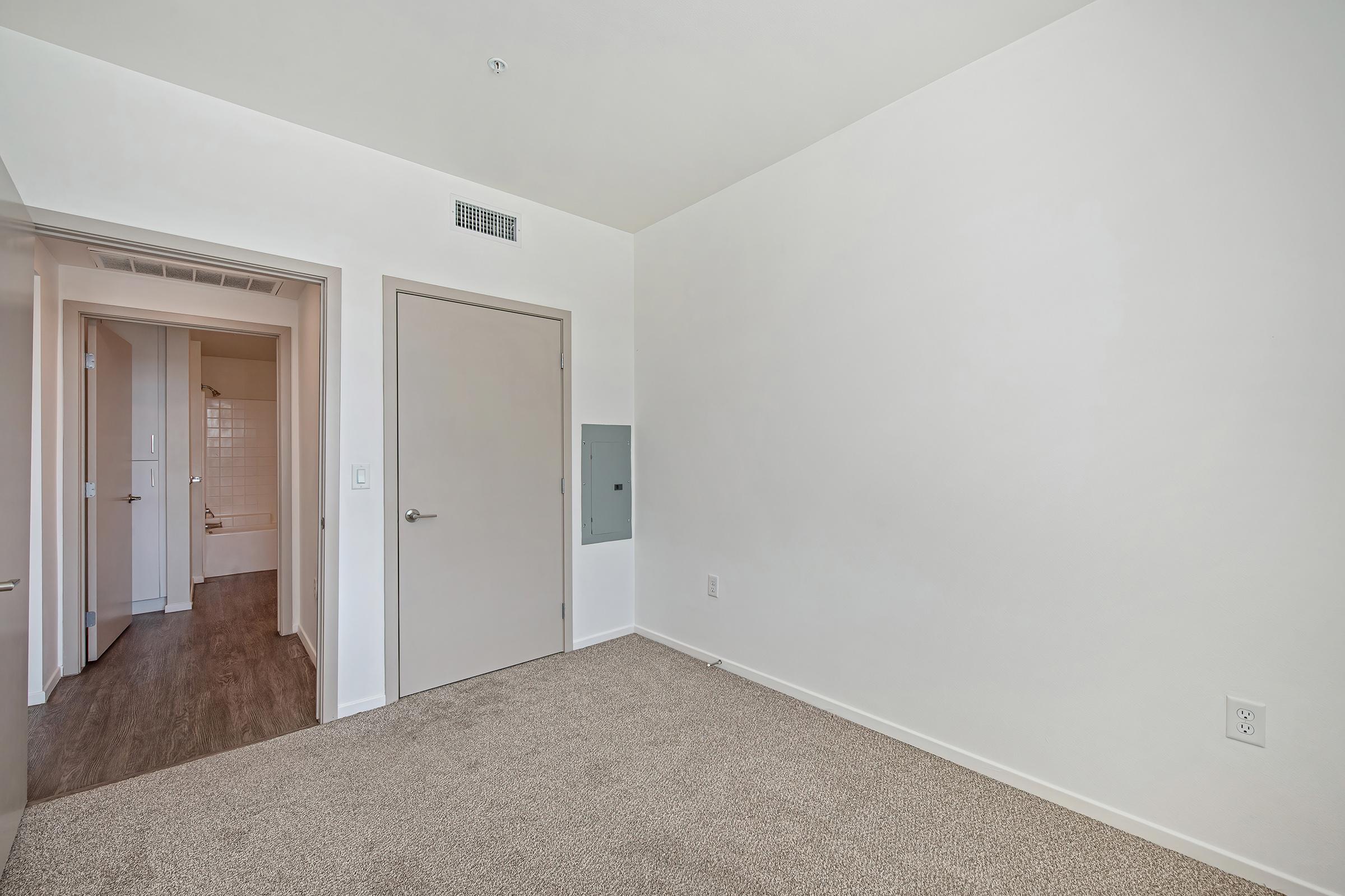 A minimalist, empty room with light-colored walls and carpet. It features a closed door leading to another room, a wall-mounted electrical panel, and an open doorway visible in the background. Natural light may be coming from off-screen, giving the space a bright, airy feel.