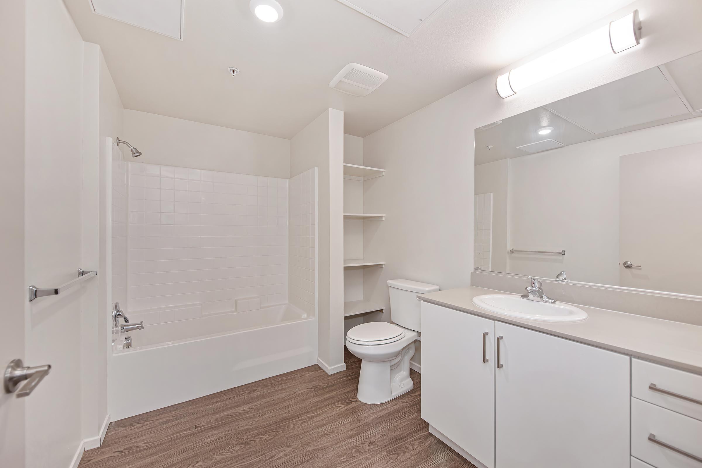 A clean, modern bathroom featuring a white bathtub with a shower, a toilet, a double sink vanity, and a large mirror. The walls are light-colored, and there is a small shelf next to the bathtub. The floor is a wood-like laminate, enhancing the aesthetic of the space. Bright lighting illuminates the area.