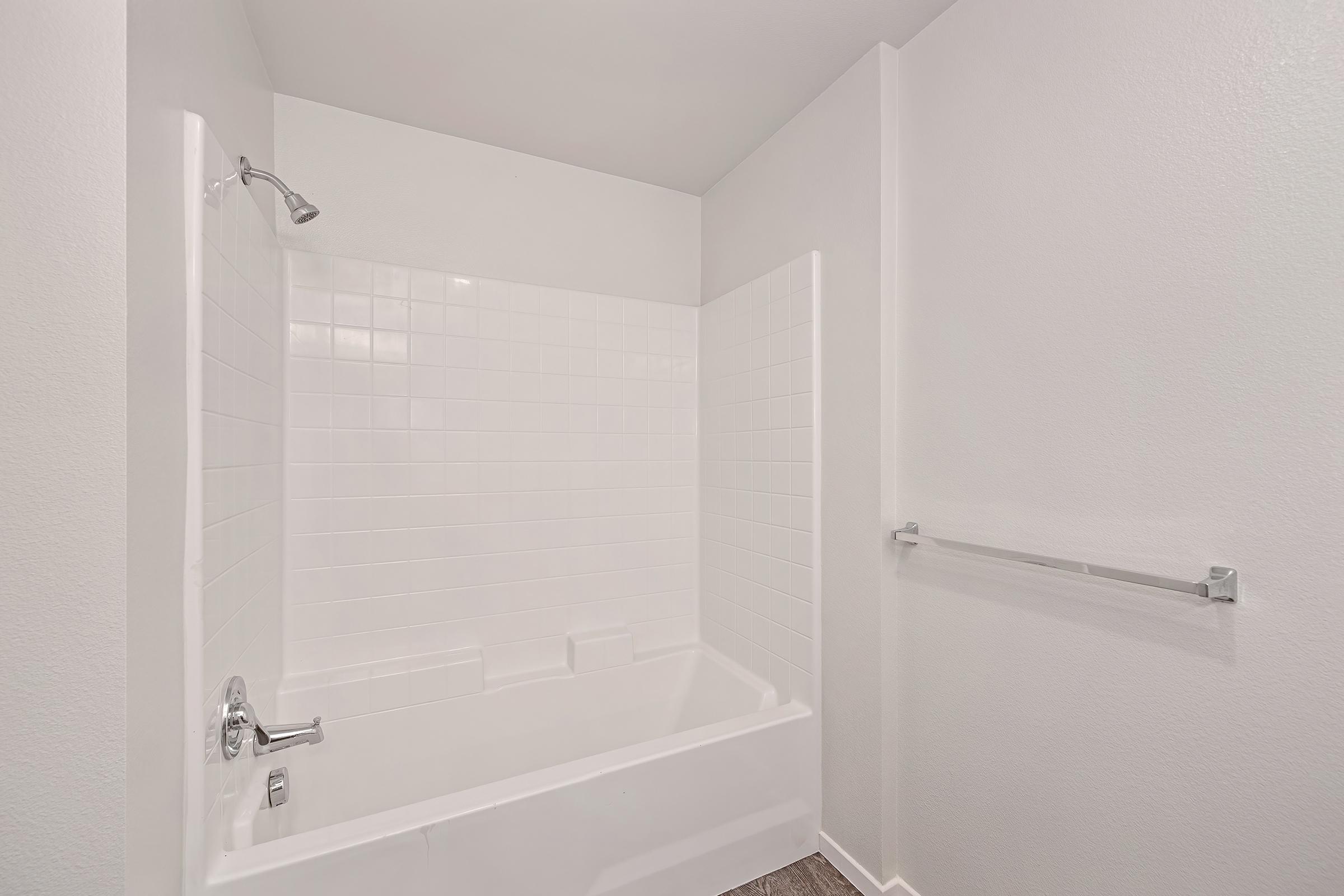 A clean, empty bathroom featuring a white bathtub with a showerhead, surrounded by glossy white tiles. The walls are painted in a light color, and there is a chrome towel bar on one side, creating a simple and modern aesthetic. The flooring is a contrasting dark shade.