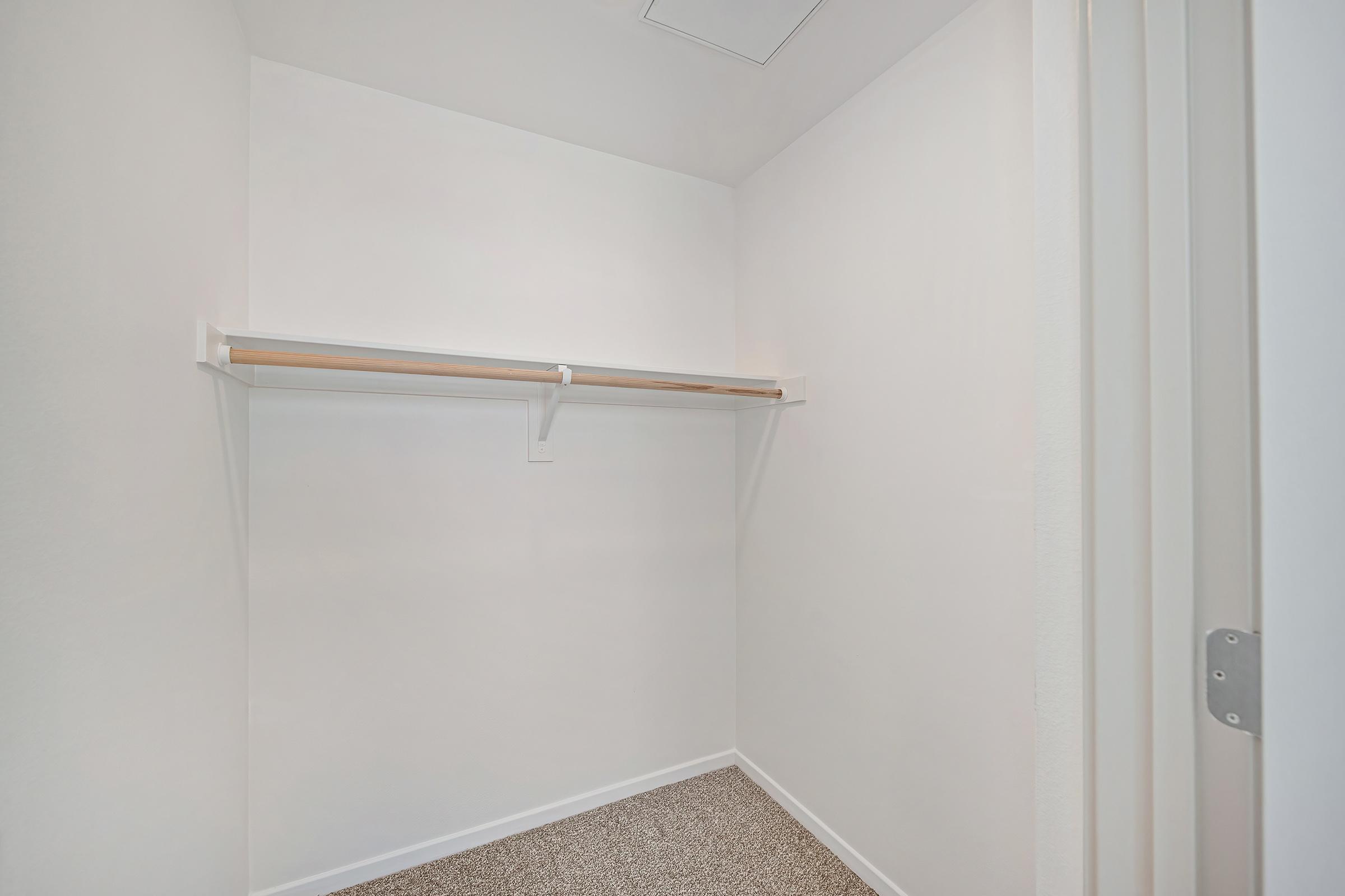 An empty closet with white walls and a light-colored carpet. There is a single wooden shelf mounted on the wall, providing a minimalist storage solution. The closet has a door slightly ajar, showing the simple and spacious interior.