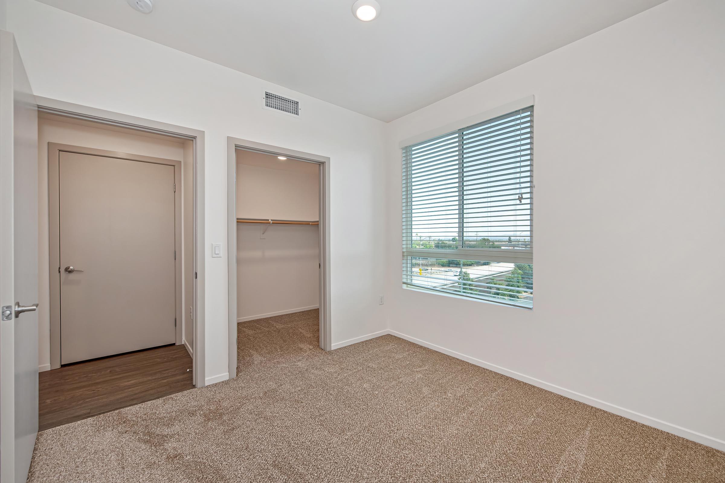 A clean, empty bedroom with light beige carpet and white walls. There is a window with horizontal blinds allowing natural light, and a partially open door leading to a closet. Another door is visible, possibly leading to a bathroom or hallway. The space is well-lit and modern, with a simple layout.