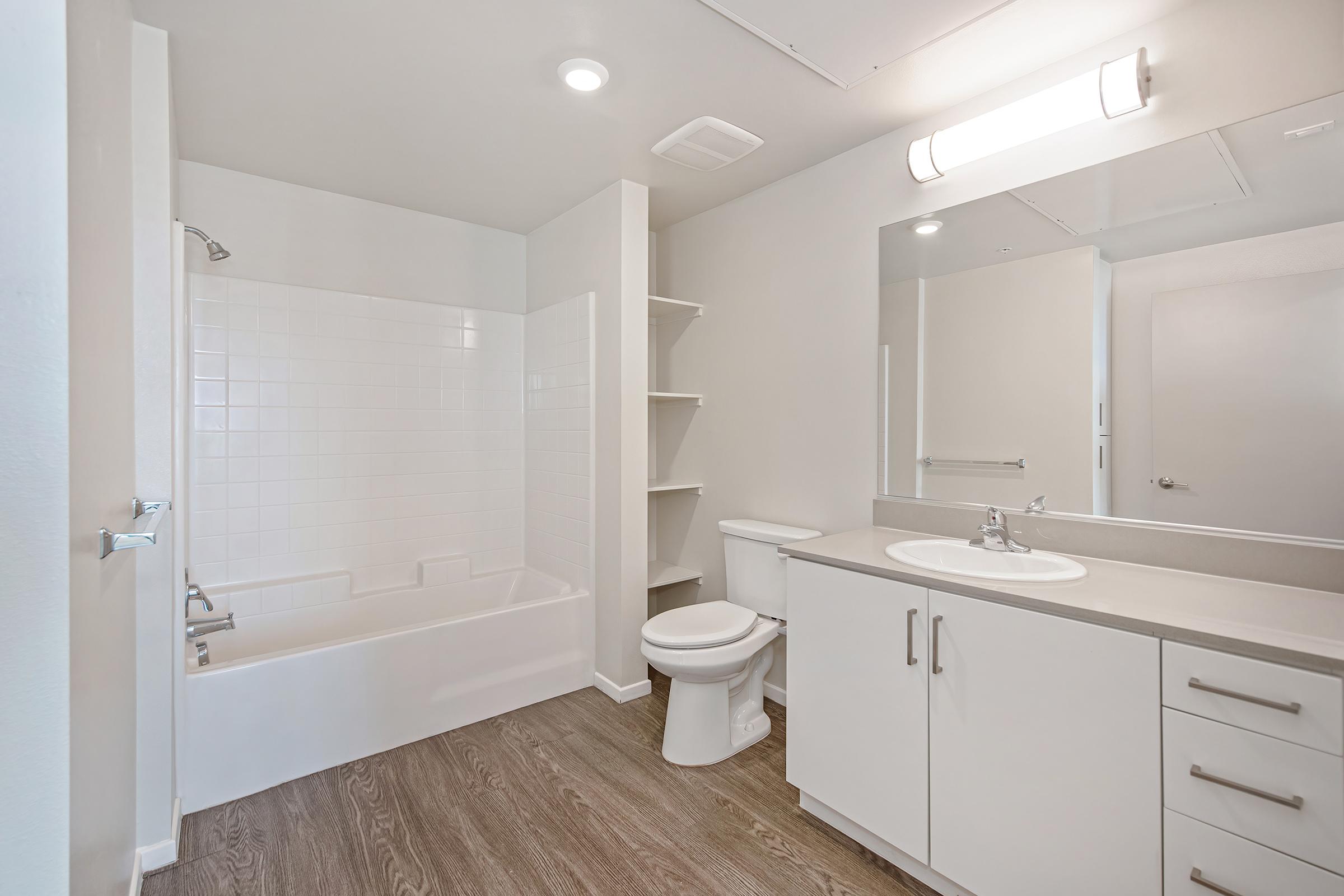 A modern bathroom featuring a white bathtub with a shower, a toilet, and a double sink vanity. The walls are light-colored, and there is a large mirror above the sink. Shelving is present beside the bathtub, and the flooring is wood-like in appearance. Bright lighting enhances the spacious feel of the room.