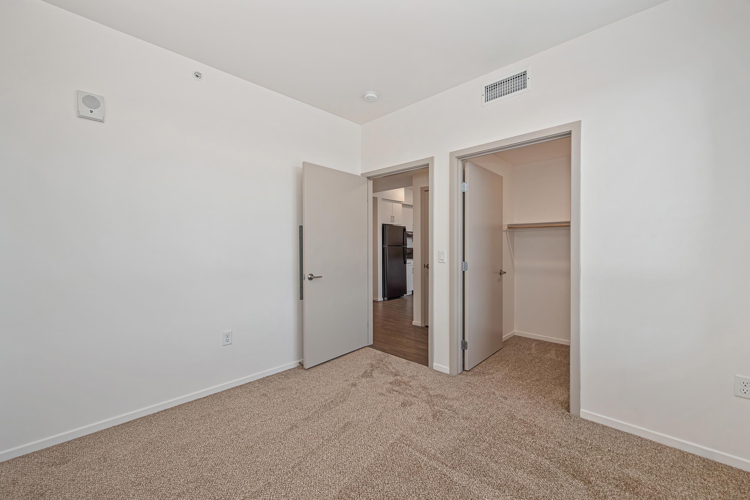 A vacant room with light-colored walls and carpet. Two doors lead to adjacent spaces—one opening to another room, and the other to a closet. A small speaker is mounted on the wall near the ceiling, and the floor features a warm-toned wooden area visible through the open door on the left.