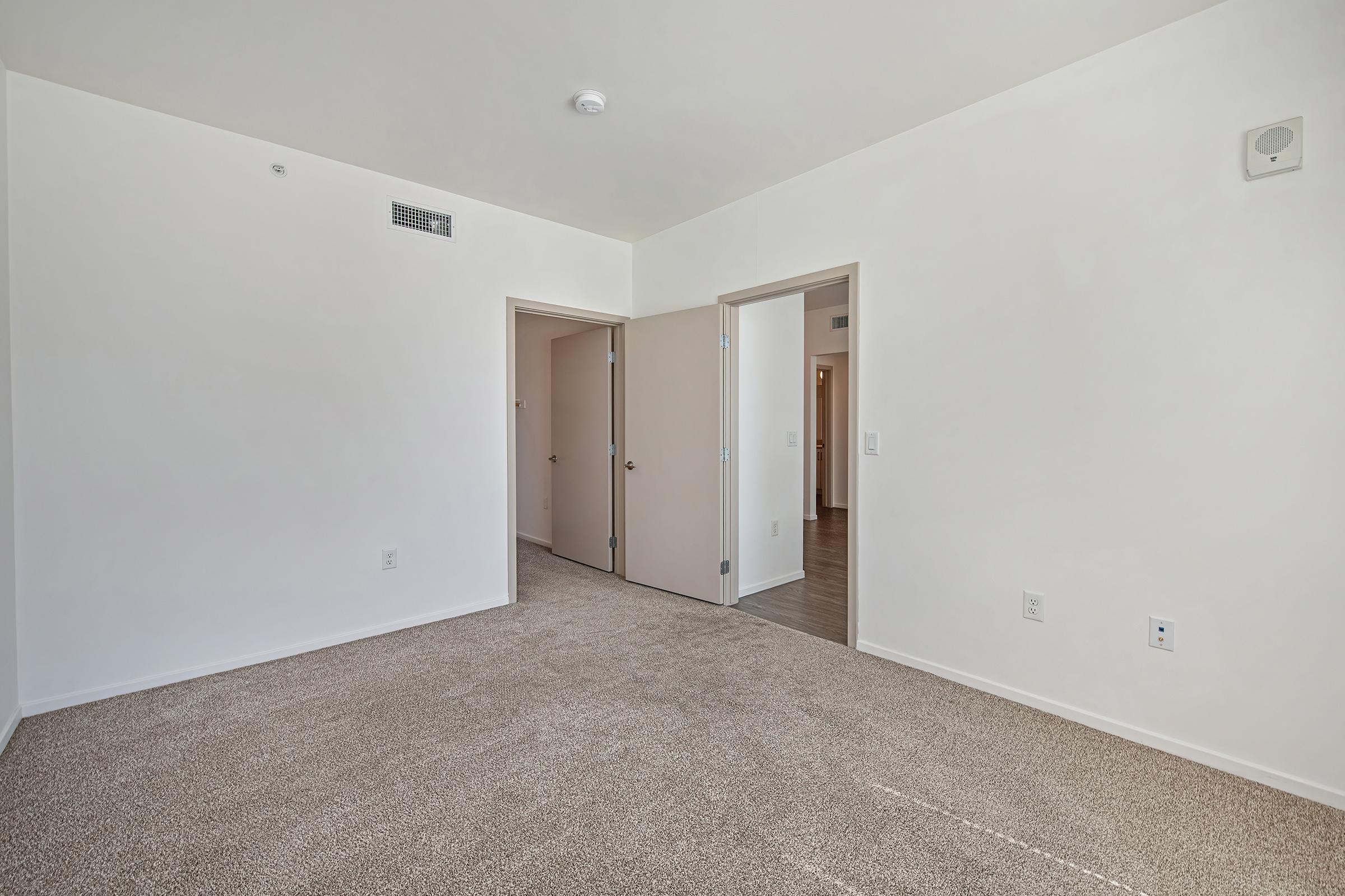 A spacious, empty room with light beige carpet and white walls. Two doorways are visible, leading to adjacent rooms, with a small wall vent and a ceiling light fixture. The room is well-lit, giving a bright and airy feel. The overall aesthetic is clean and minimalistic.