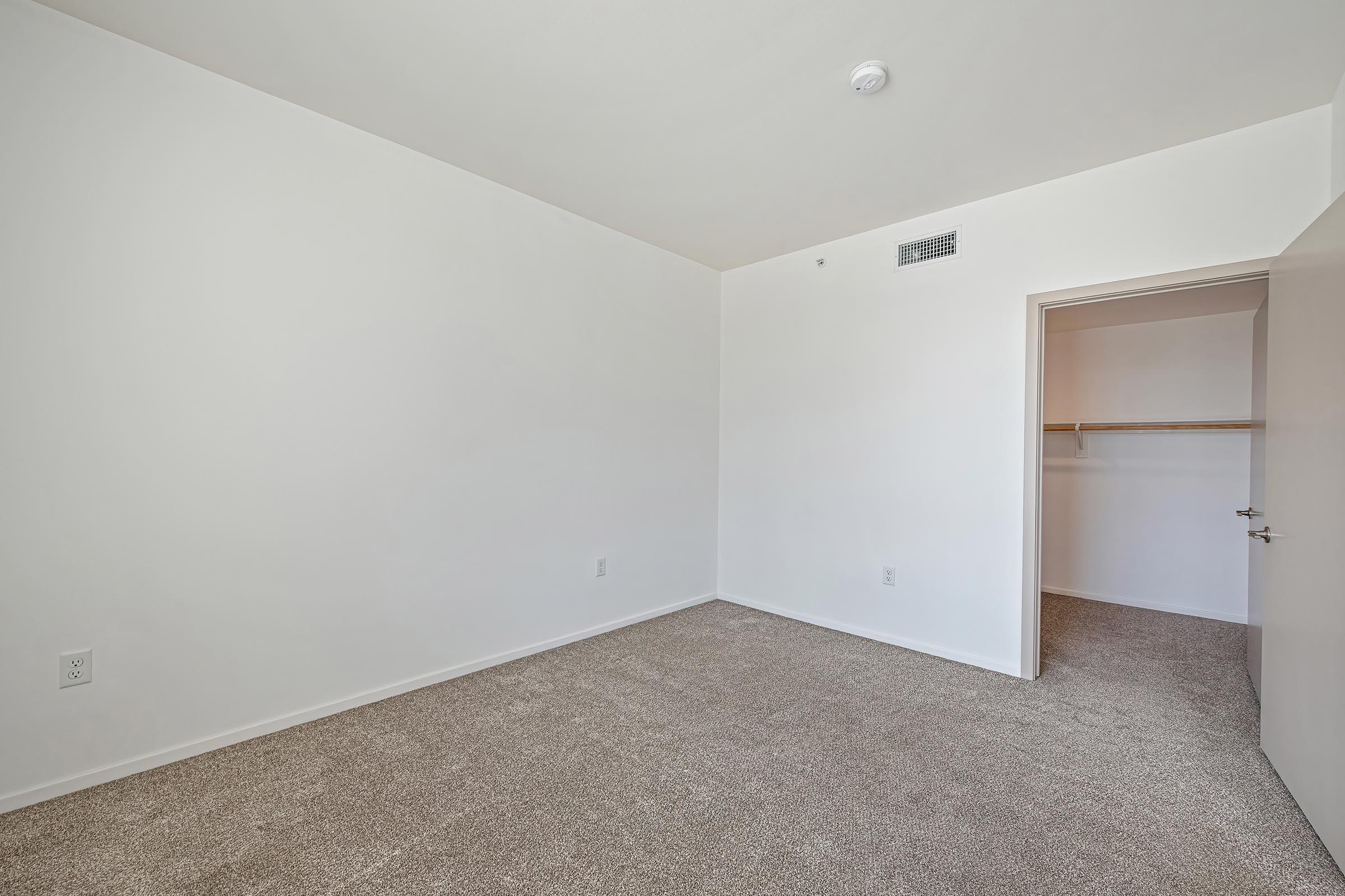 A vacant room with beige carpet and white walls. There is a closet with a shelf visible through an open door on the right. The room has a single ceiling light fixture and no furniture or decorations, creating a minimalistic and spacious appearance.