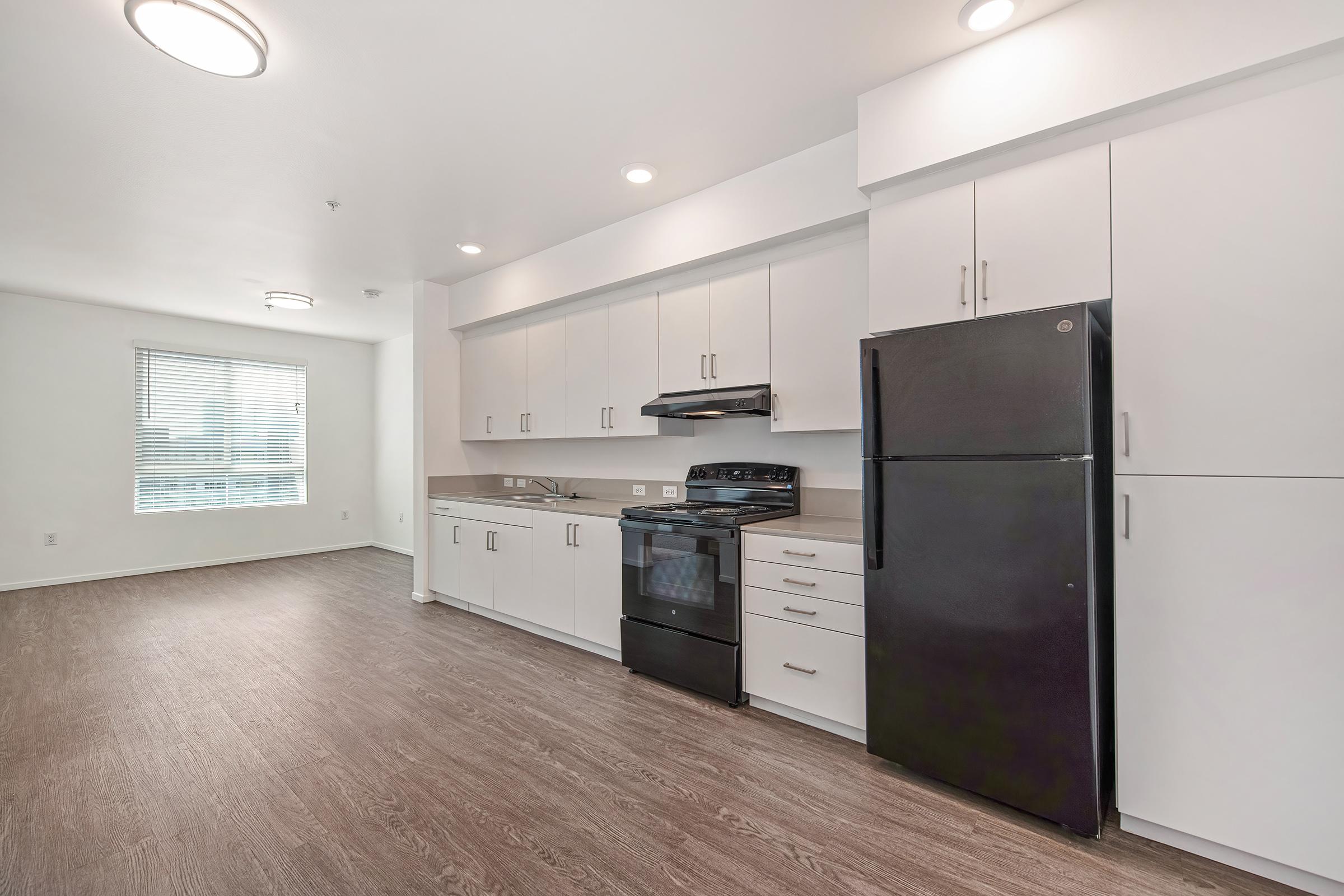 Modern kitchen featuring white cabinetry, a black refrigerator, and a stainless steel stove. The open layout includes a large window letting in natural light, with a neutral-colored floor and minimalistic design elements. Bright overhead lighting enhances the spacious feel of the room.