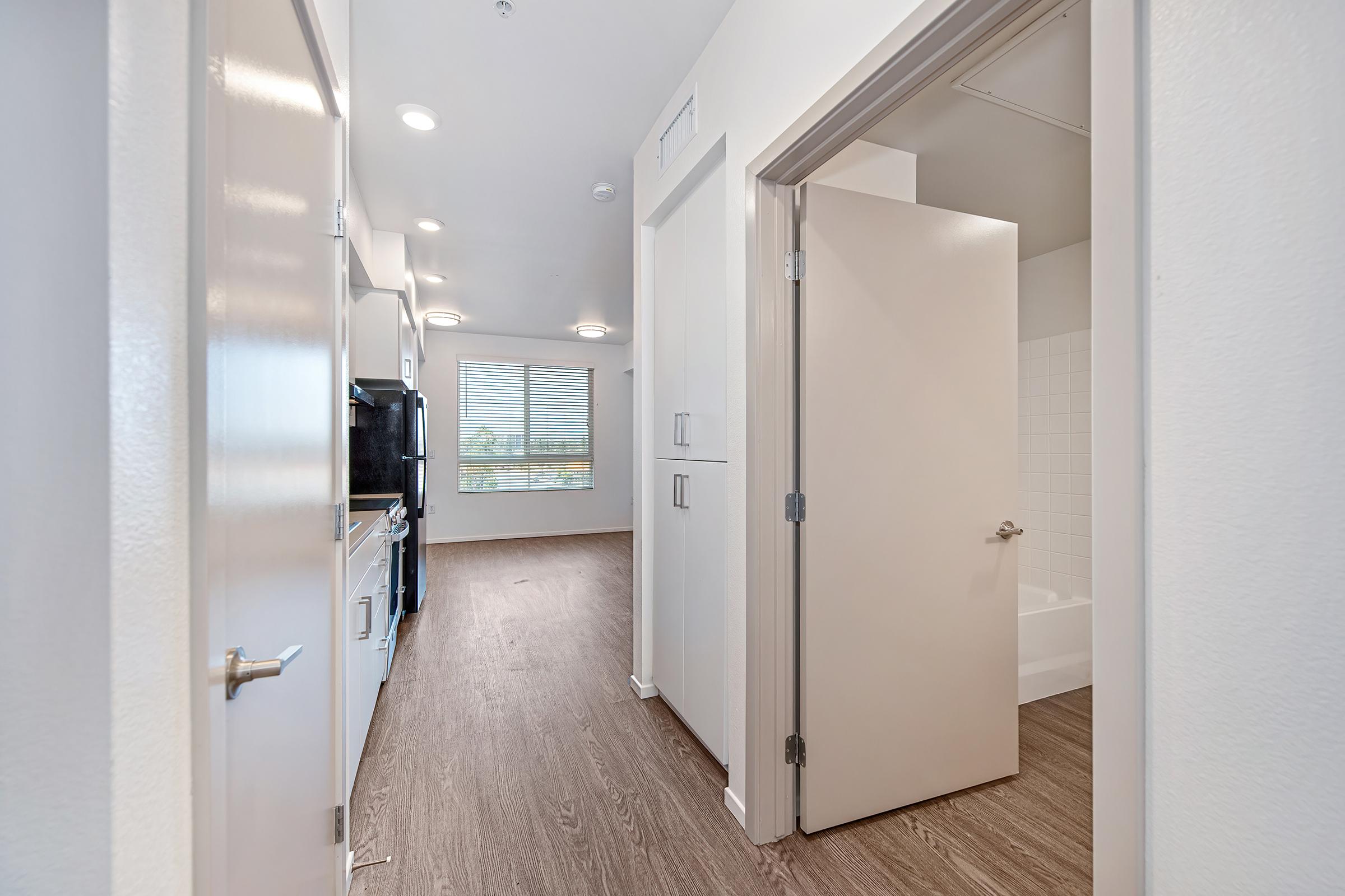 A well-lit interior view of an apartment, featuring a clean kitchen area on the left with modern appliances, a hallway leading to a bathroom on the right, and a window with blinds offering natural light. The flooring is a sleek, light-colored laminate, creating a spacious and inviting atmosphere.