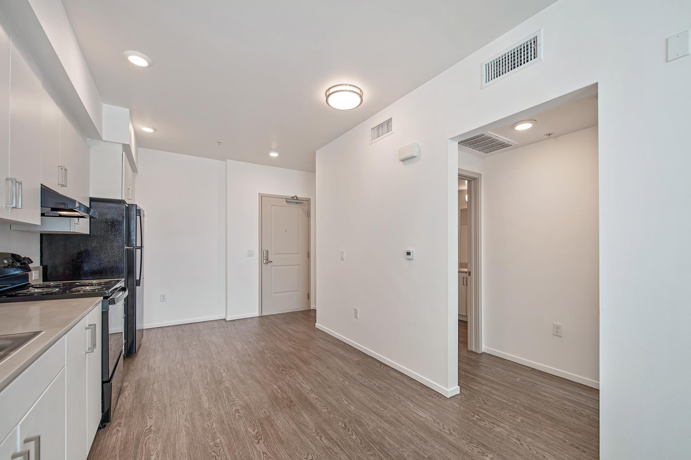 A modern kitchen and living area featuring white cabinets, stainless steel appliances, and wood-style flooring. An open doorway leads to a hallway, with a light-colored wall and a door visible in the background. Ceiling lights provide bright illumination in the spacious, uncluttered environment.