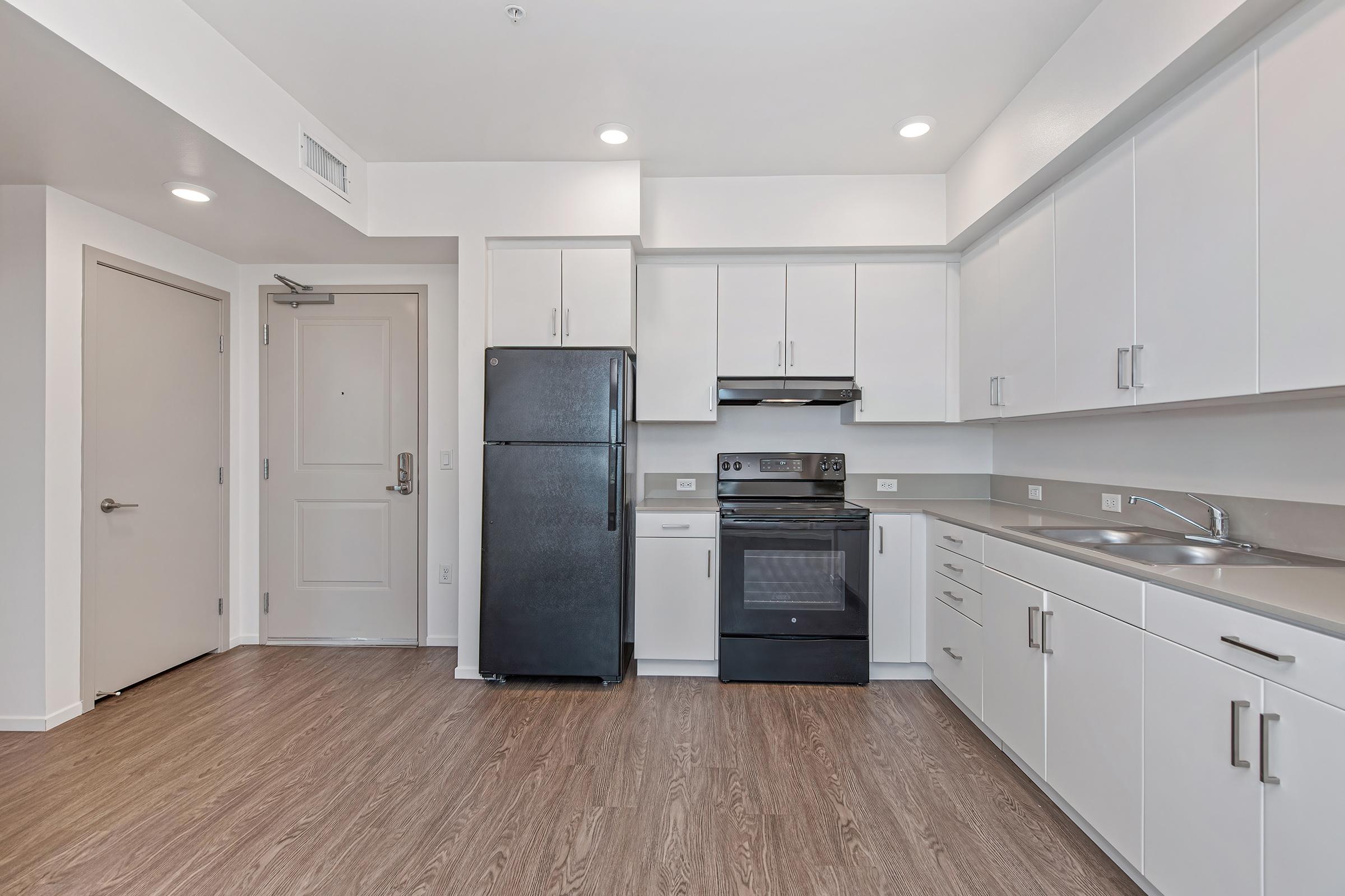 Modern kitchen featuring a black refrigerator and stove, white cabinetry, and a stainless steel sink. The space has light-colored walls and a wood-like floor, with a door leading out and ample overhead lighting.