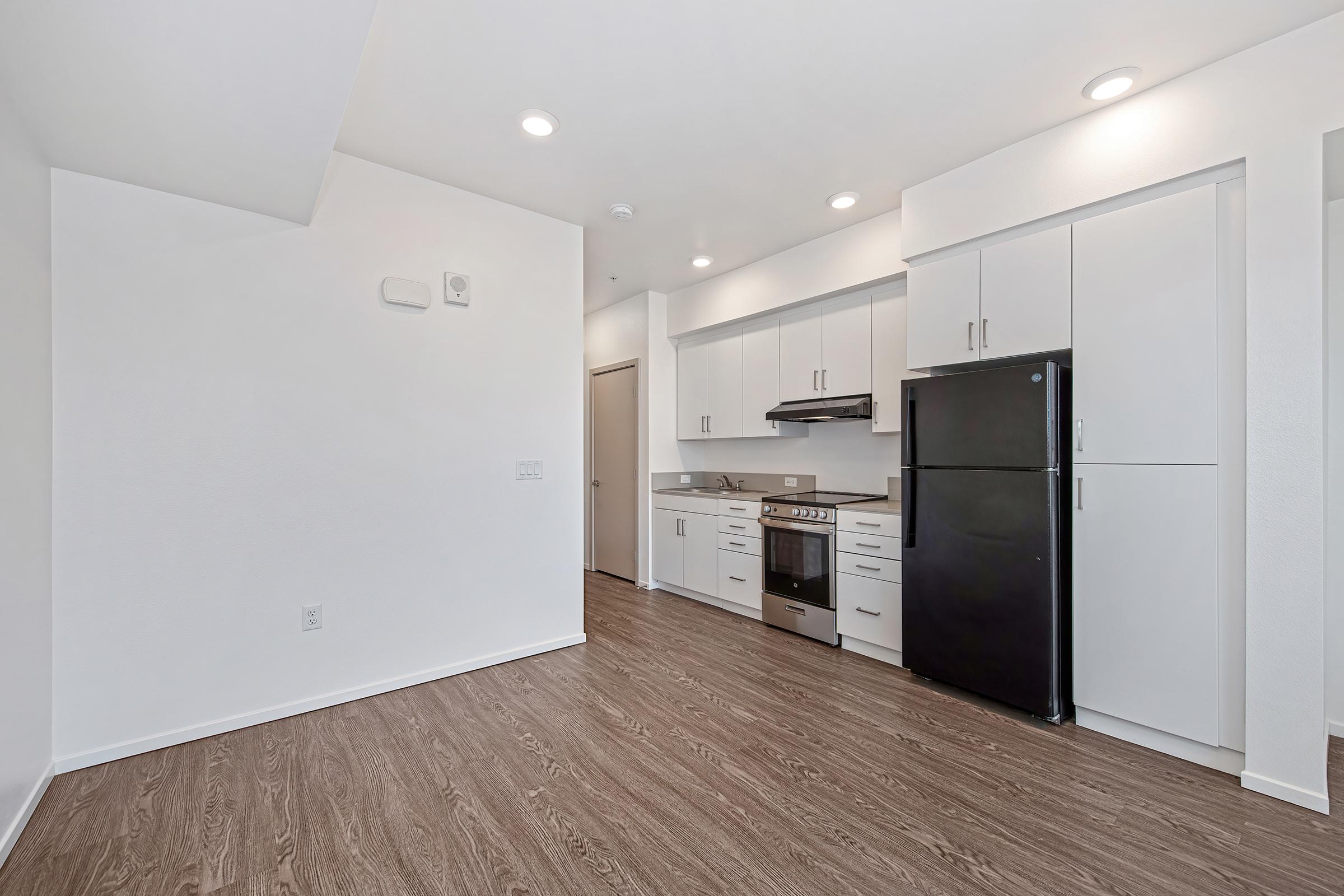 A modern kitchen in an apartment featuring white cabinets, a black refrigerator, and stainless steel appliances. The space has an open layout with wood-style flooring and bright lighting, creating a clean and contemporary look.