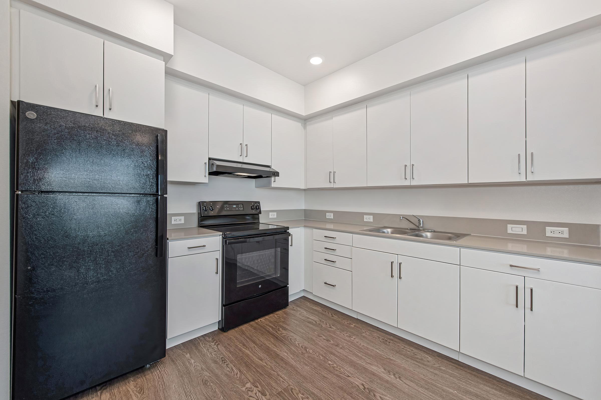 A modern kitchen featuring a black refrigerator, black stove, and white cabinetry. The countertops are light-colored, and the floor is made of wood. Bright lighting illuminates the space, creating a clean and contemporary atmosphere.