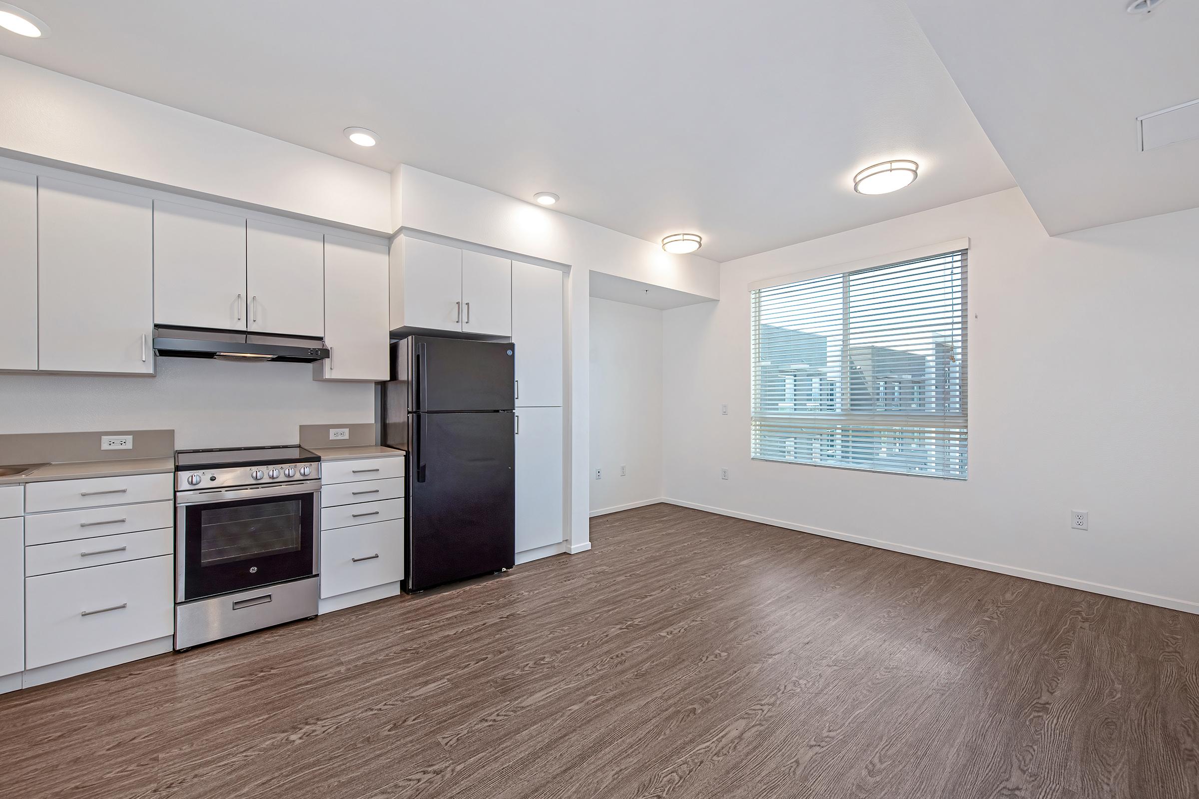 Modern kitchen with white cabinets, a black refrigerator, and a stainless steel oven. There is an overhead microwave and a large window with blinds, allowing natural light to fill the space. The floor is wood-like laminate, creating a warm atmosphere in the open layout.