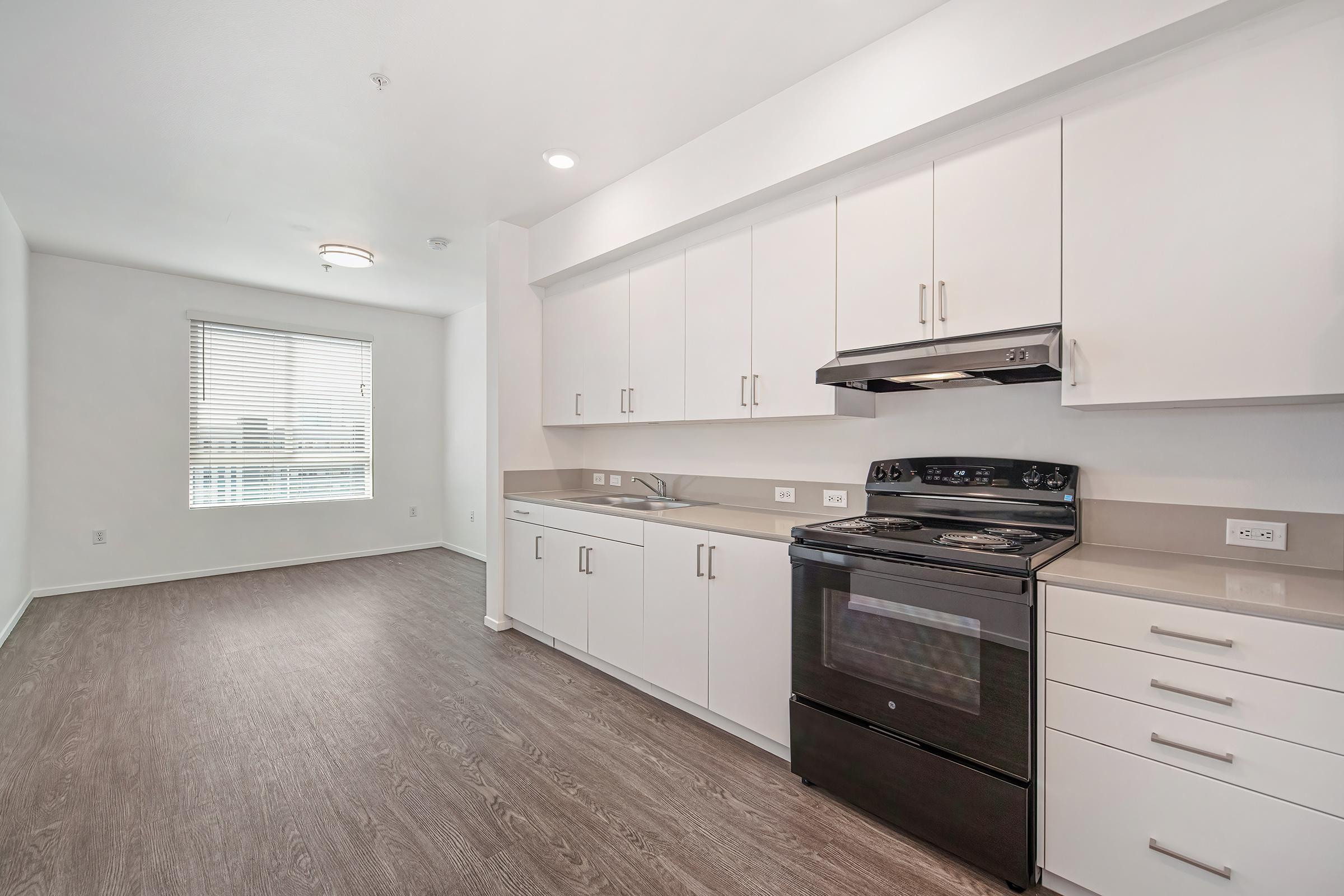 A modern kitchen featuring white cabinets, a black stove, and a spacious countertop. Natural light enters through a window with blinds, illuminating the clean and minimalistic design. The flooring is a light-colored wood laminate, adding warmth to the overall aesthetic.